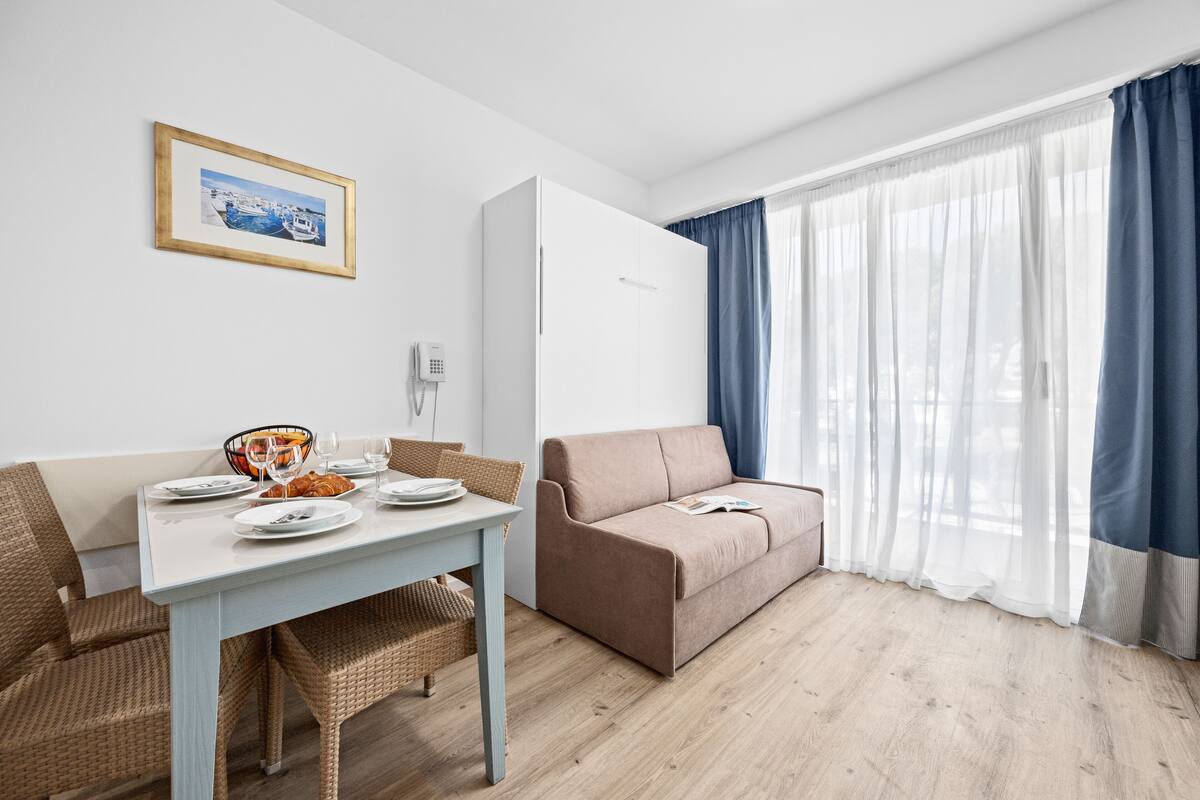 Una sala moderna con una mesa de comedor preparada para dos personas, un sofá y un ventanal con cortinas.