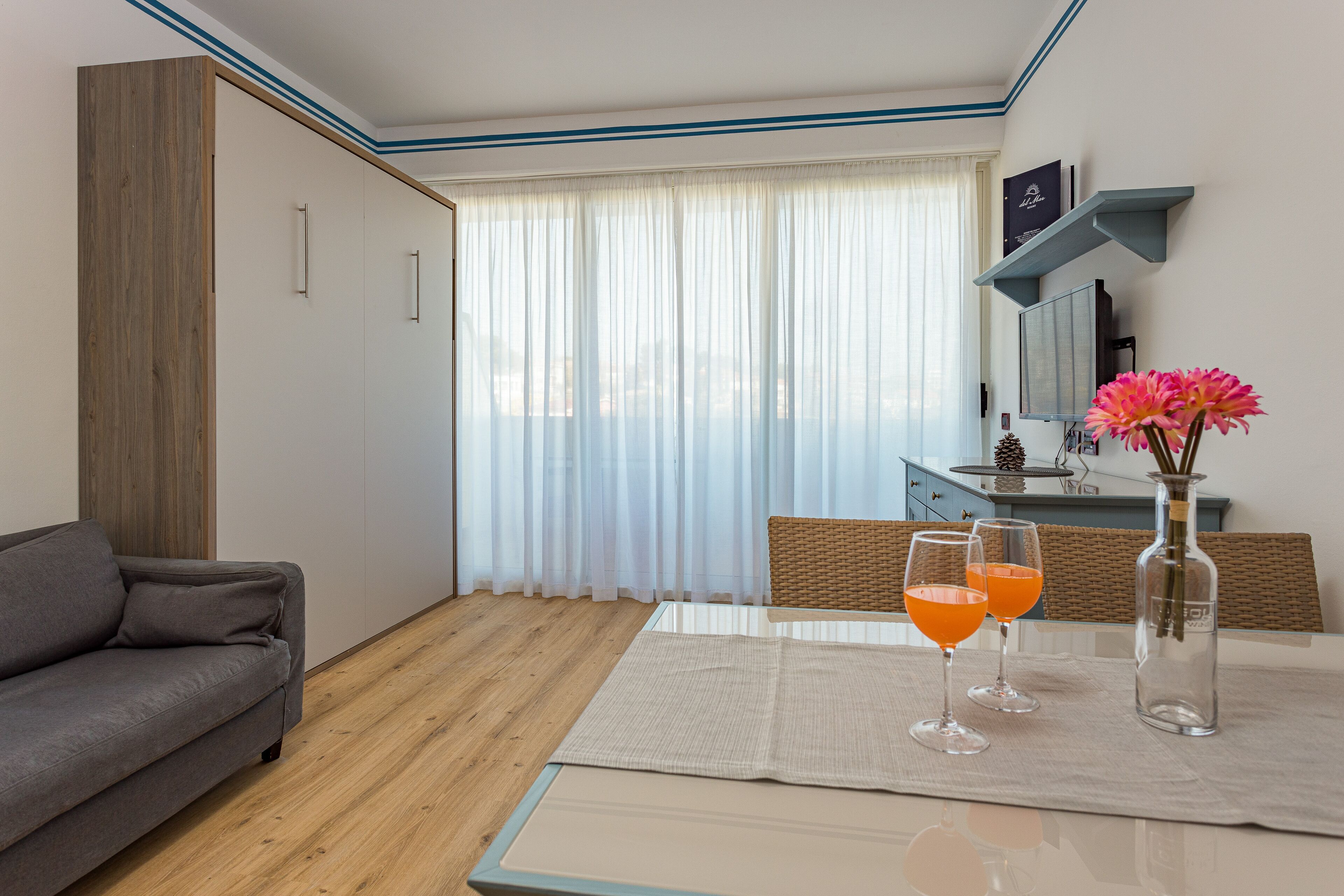 Une chambre moderne avec un canapé, une table à manger sur laquelle sont posés deux verres de jus d’orange, un vase rempli de fleurs et une armoire en bois.