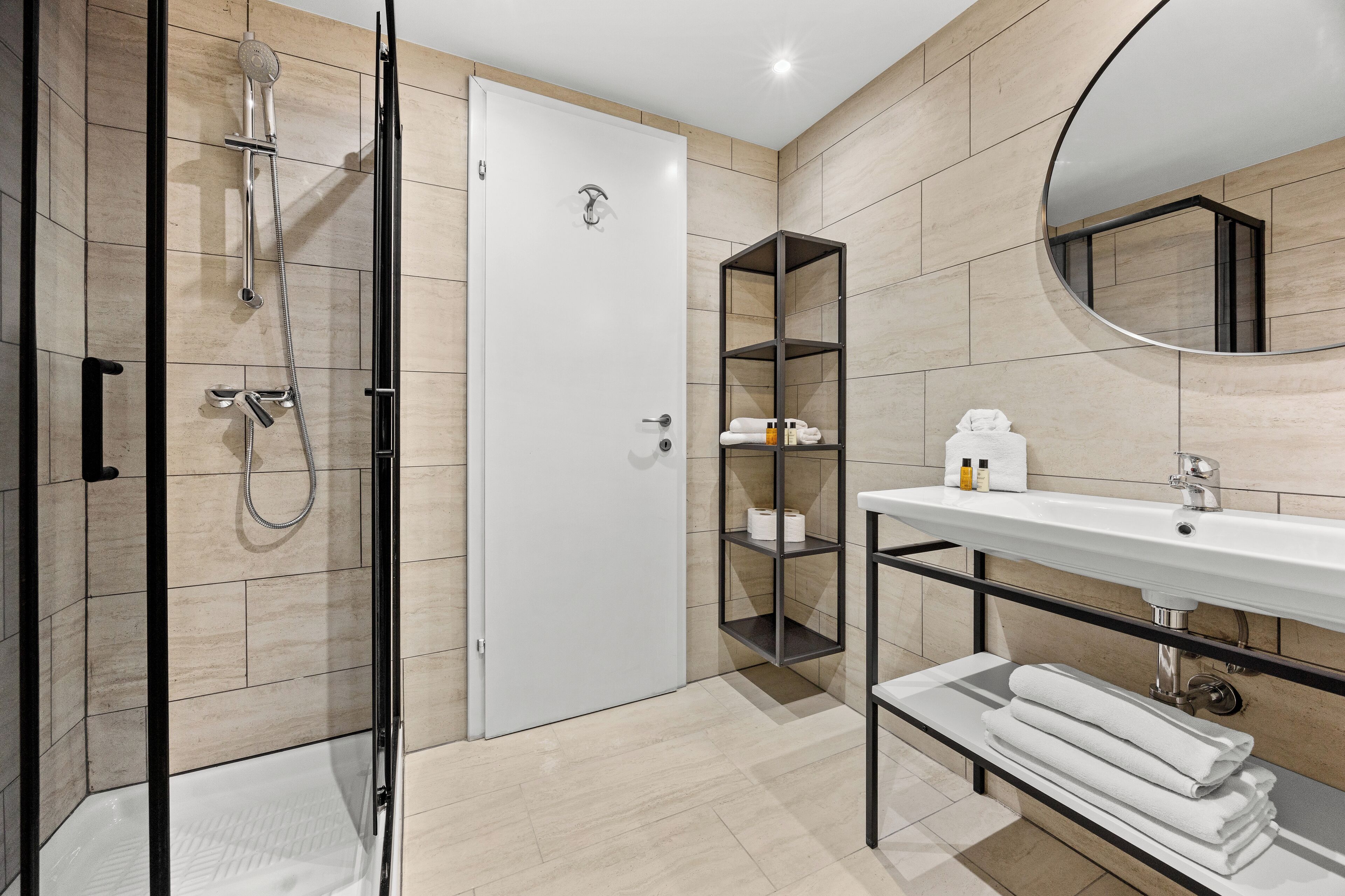 Een moderne badkamer met een glazen douchecabine, een witte deur, een wastafel met spiegel en planken met toiletartikelen.