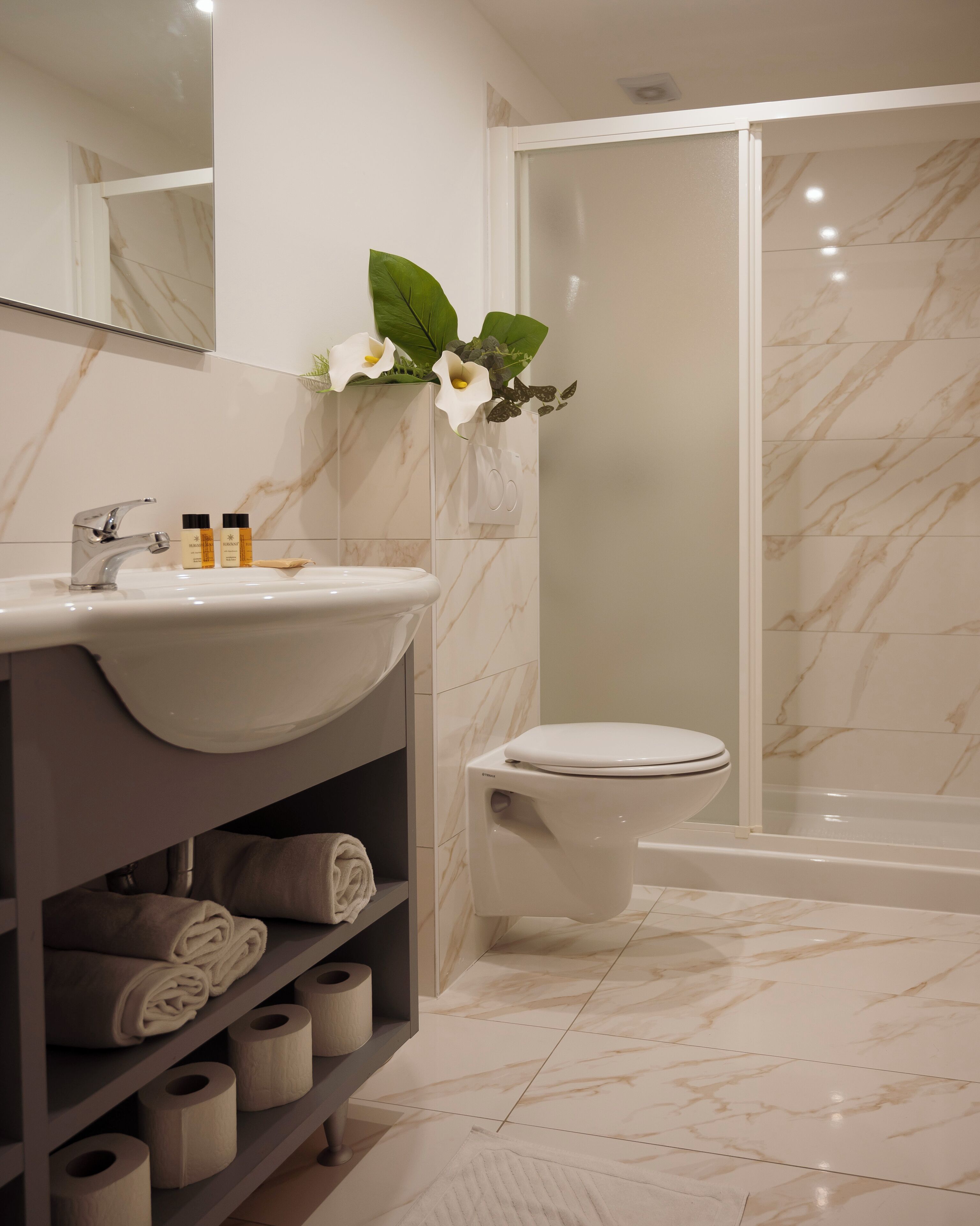 Un baño moderno con lavamanos, inodoro y ducha de mármol. Hay toallas y papel higiénico en estantes.