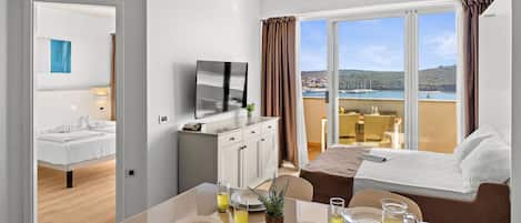 A hotel room with a bed, a dining table with breakfast items, a TV, and a balcony with a view of the sea.