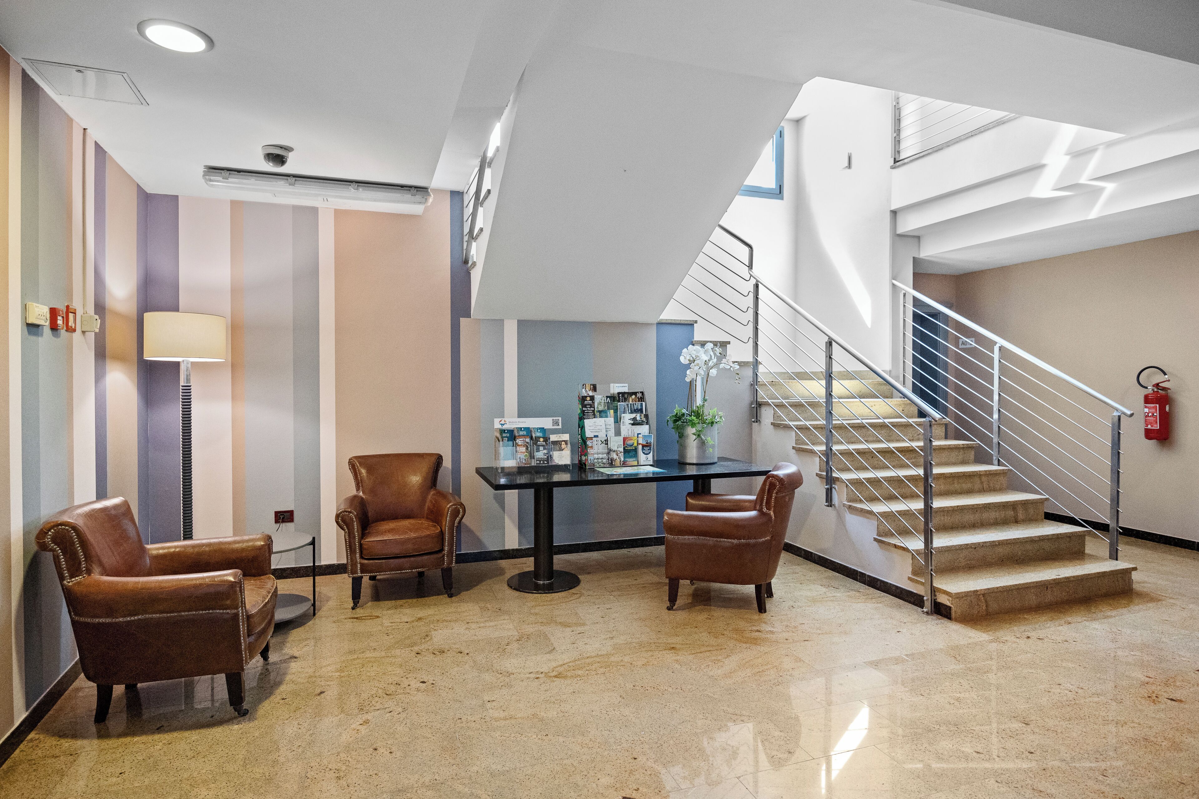Vestíbulo con escalera, sillones de cuero, una mesa con folletos y un extintor.