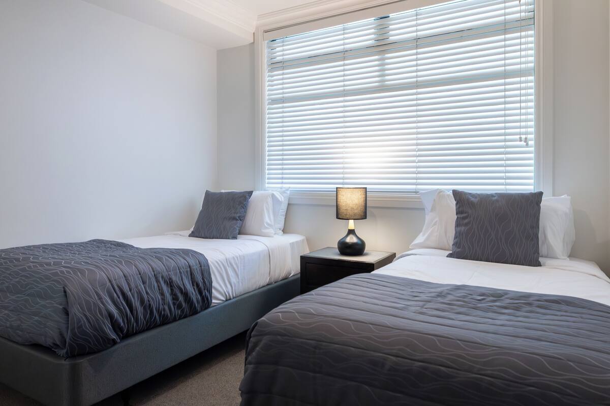 Habitación doble con dos camas, una mesita de noche con lámpara y una ventana con persianas.