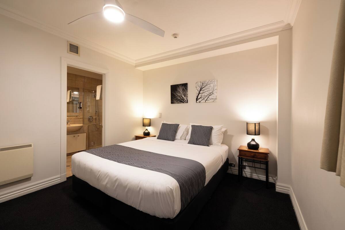 Una habitación de hotel con una cama grande, dos mesitas de noche con lámparas, una obra de arte en la pared y un baño visible a través de una puerta abierta.