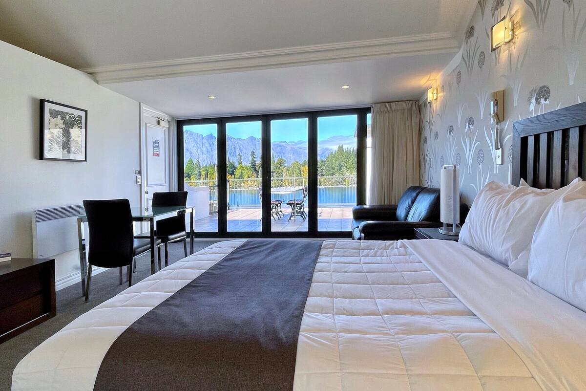 Habitación de hotel con una cama grande, un escritorio, una silla y vistas a las montañas a través de las ventanas.
