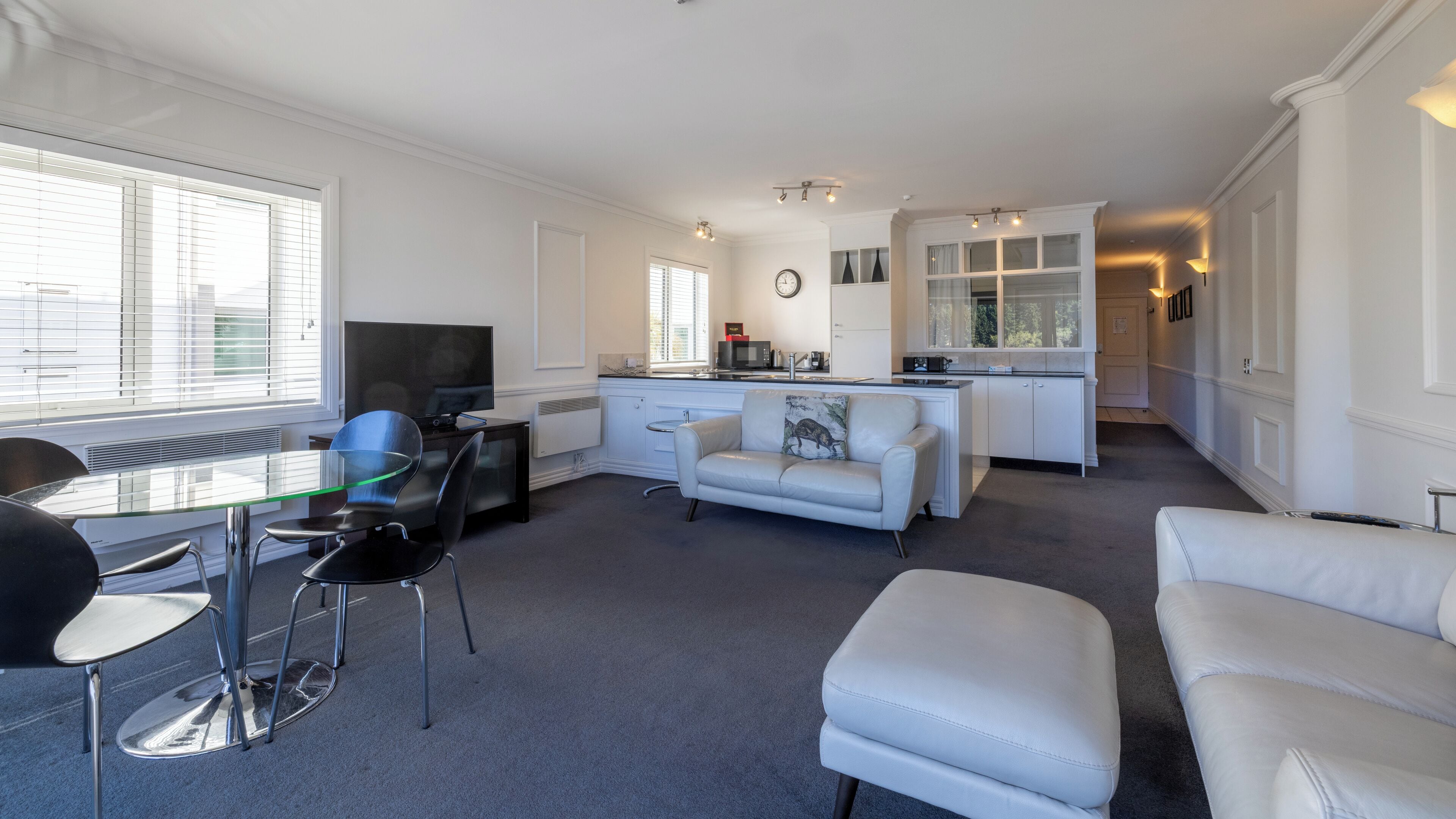 A modern living room with a glass dining table, a sofa, and a TV. There is a kitchen area with a microwave and a clock on the wall.