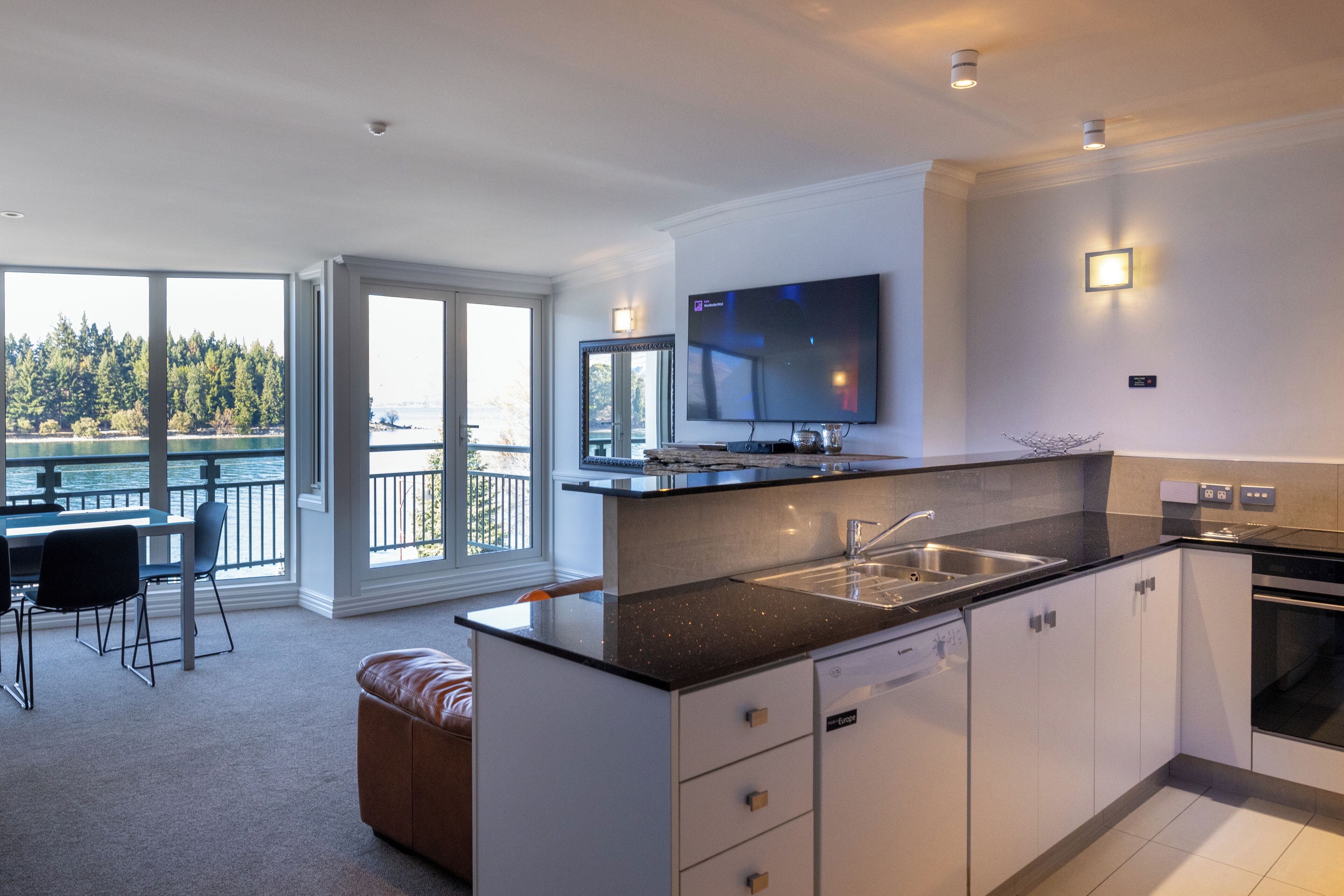 A modern kitchen with a large island, a built-in oven, and a flat-screen TV. There is a dining area with a table and chairs, and a view of the outdoors through large windows.