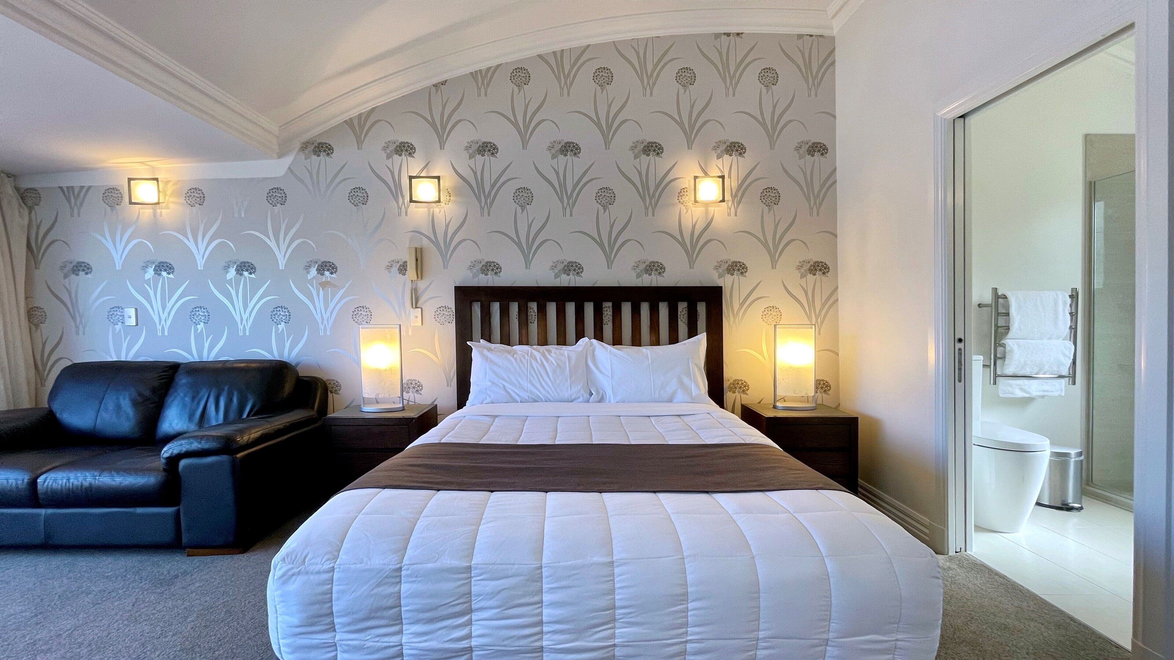A hotel room with a large bed, a dark leather sofa, two bedside tables with lamps, a wooden headboard, and a bathroom visible through an open door.