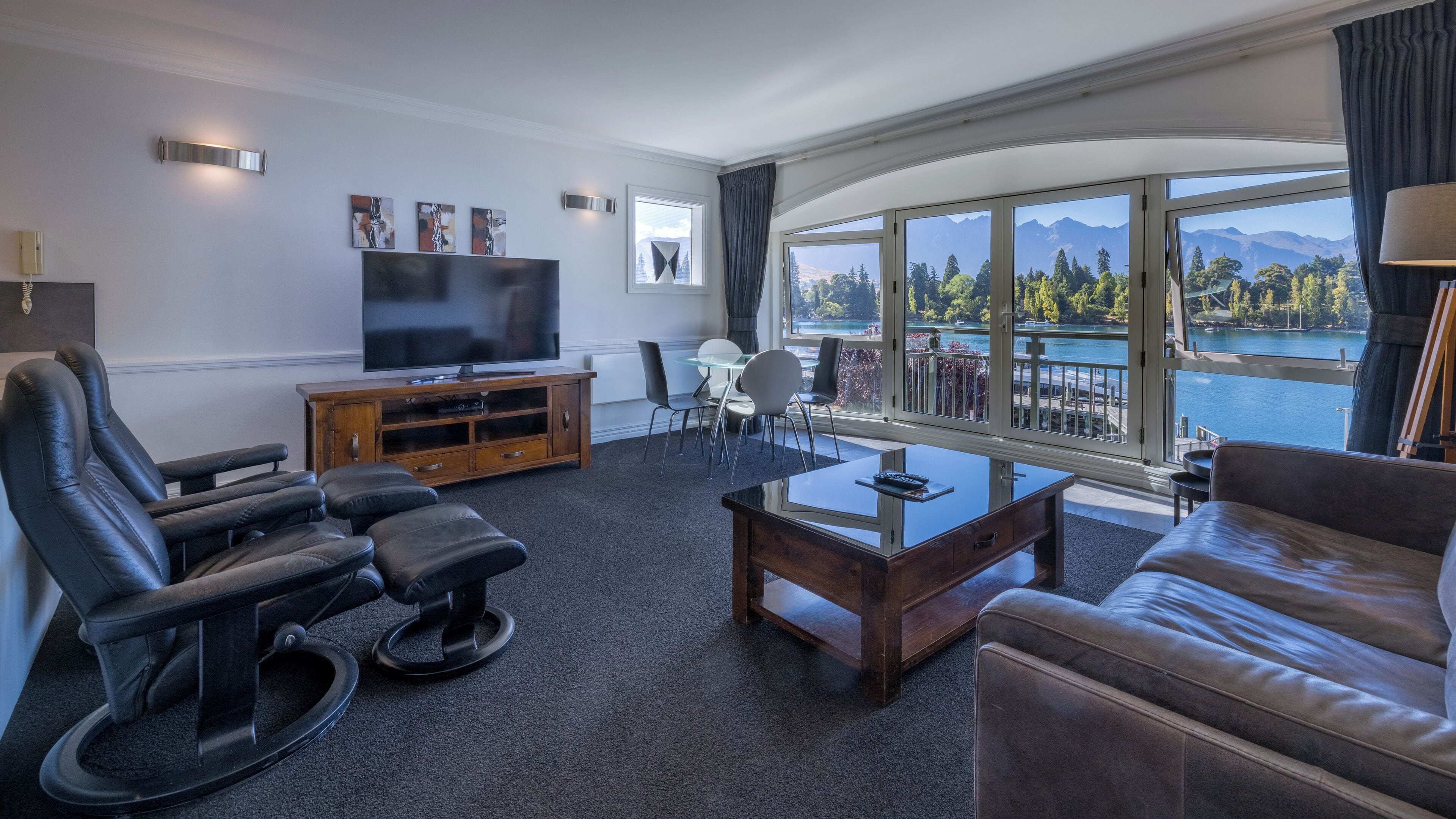 Ein modernes Wohnzimmer mit großem Flachbildfernseher, Ledersitzmöbeln, einem Glastisch und Blick auf einen See und Berge durch große Fenster.