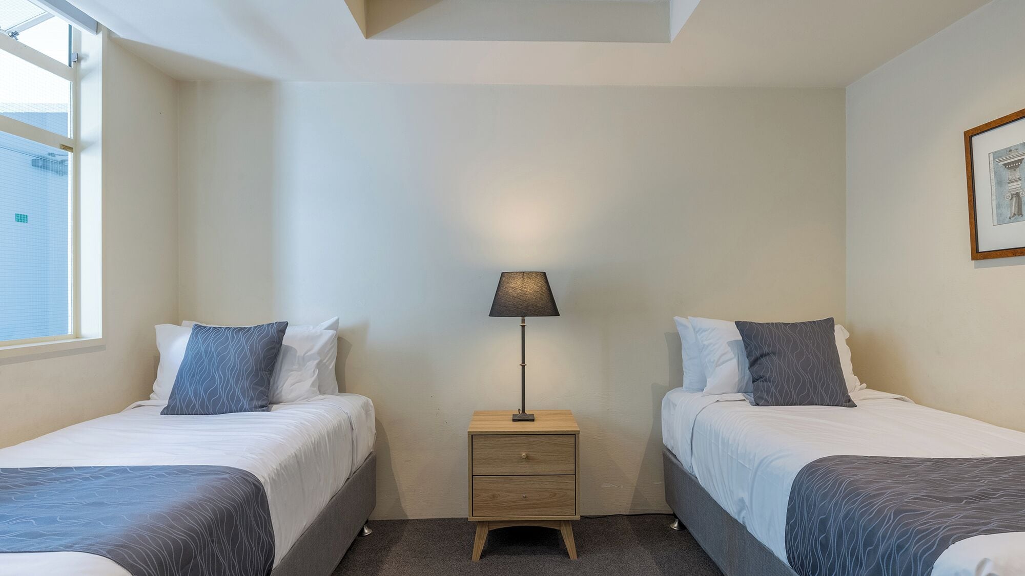 Et hotelværelse med to senge, et natbord med en lampe og et vindue.