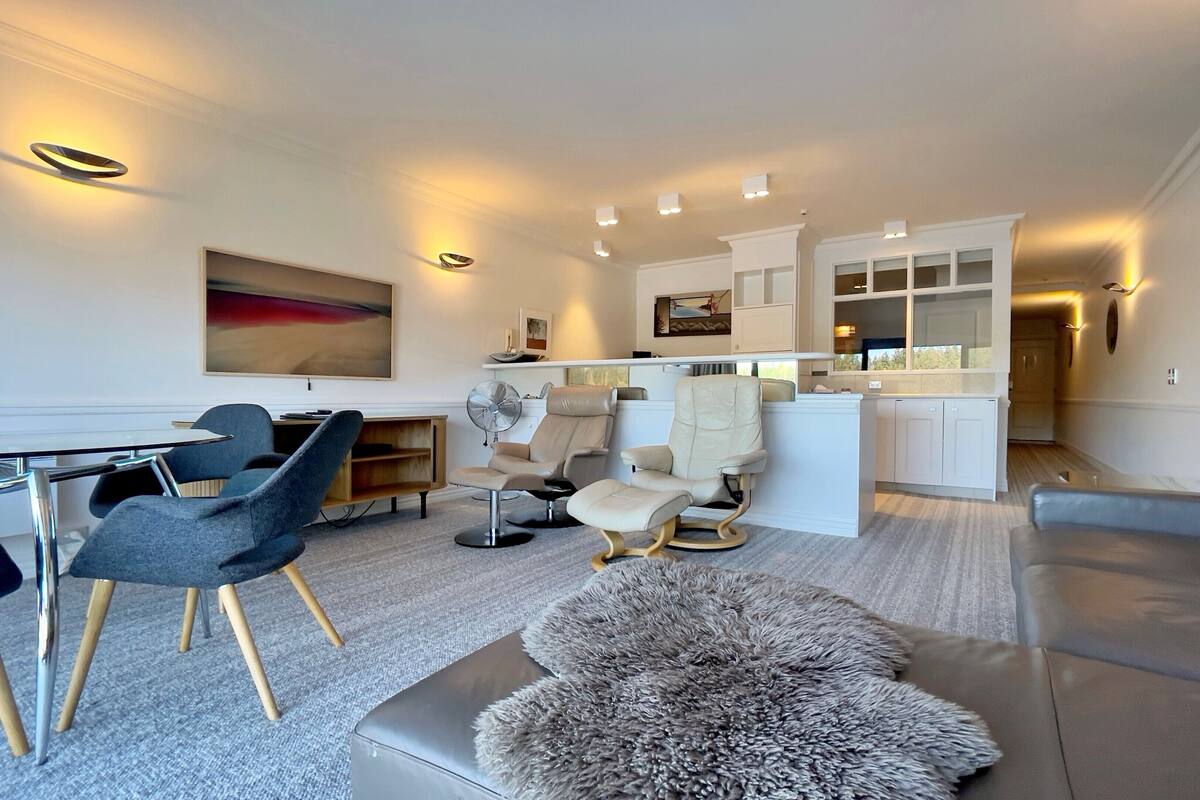 Una sala moderna con un sofá, sillas, una mesa de centro y una zona de comedor con mesa y sillas. Hay una cocina con armarios y una encimera.