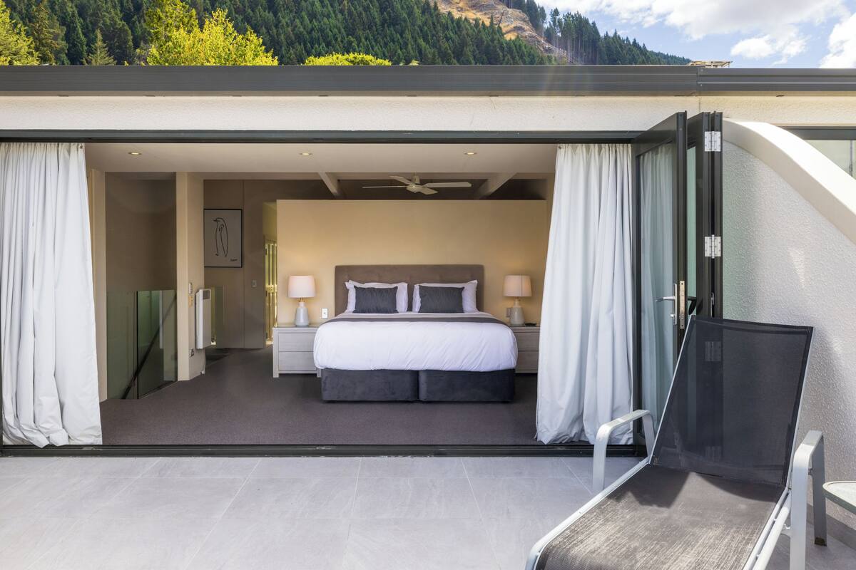 Una habitación de hotel moderna con una cama grande, dos mesitas de noche con lámparas y vistas a una montaña boscosa.