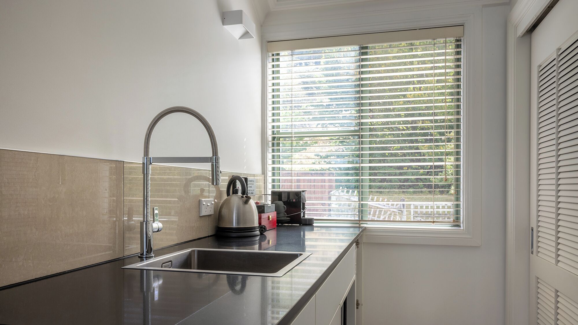 A modern kitchen with a stainless steel sink, a kettle, and a window with blinds.
