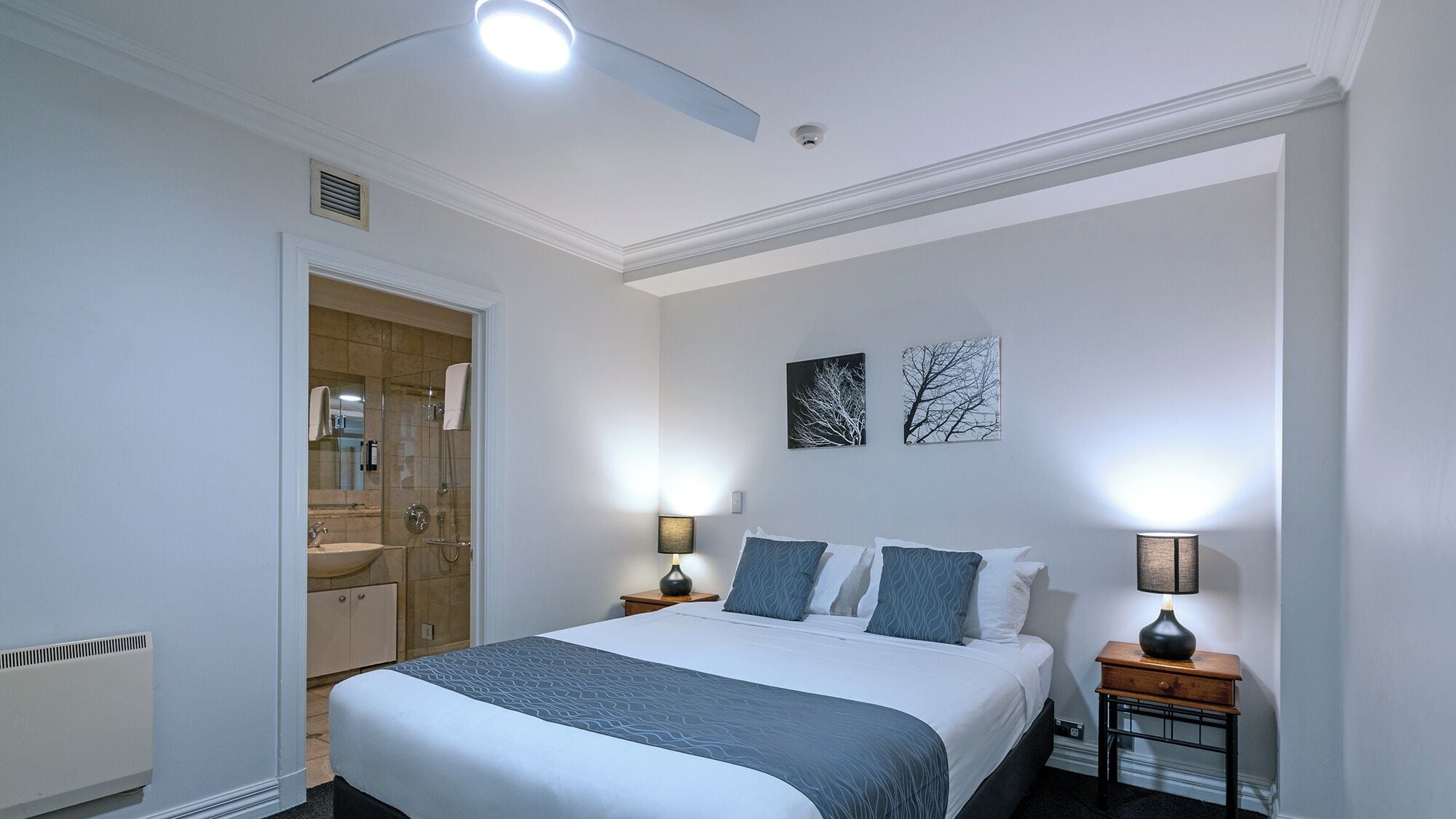 Ein Hotelzimmer mit einem großen Bett, zwei Nachttischen mit Lampen, einem Badezimmer mit Waschbecken und Spiegel sowie zwei Wandbildern.
