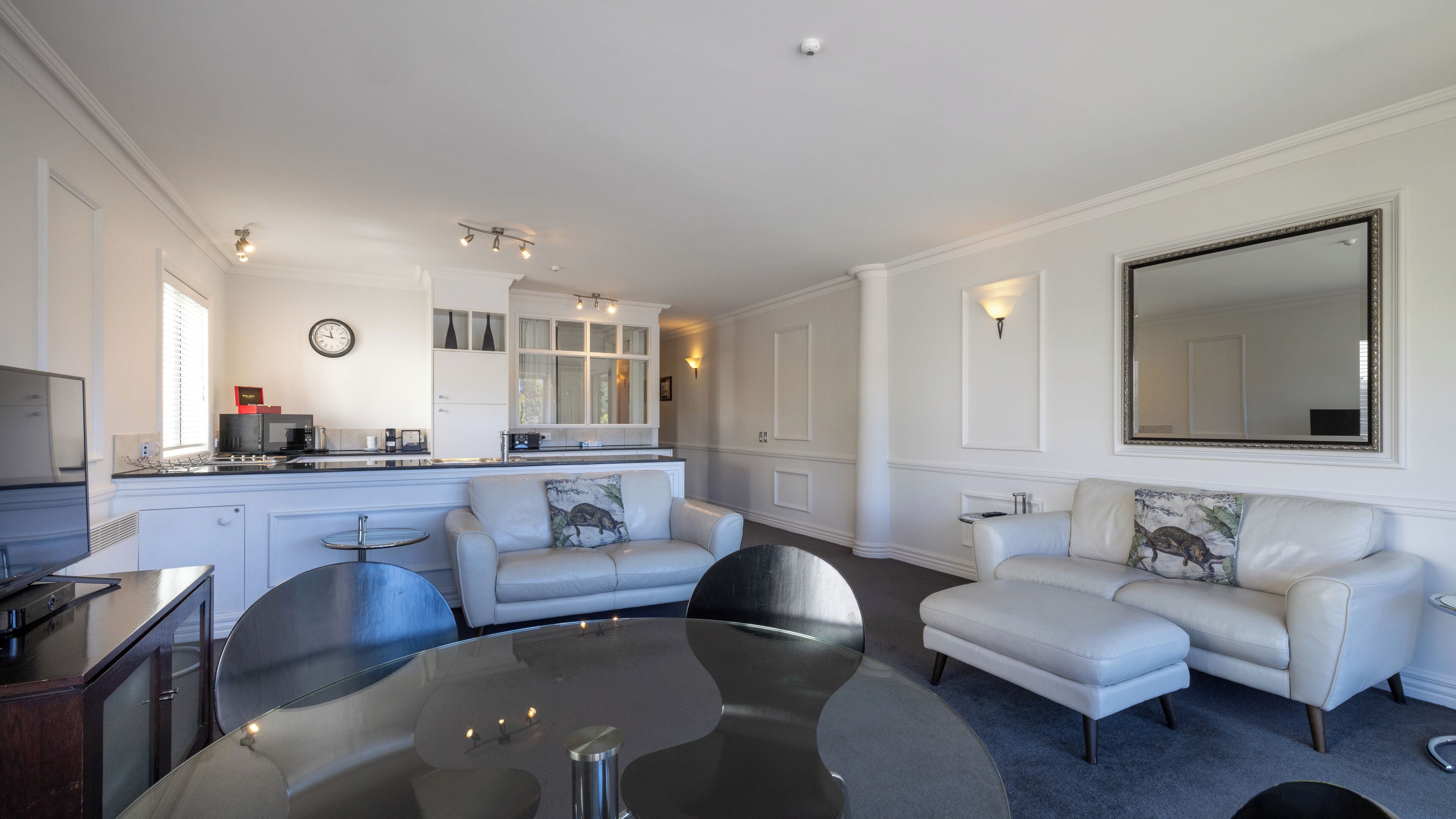 Een moderne keuken met witte kasten, een eethoek met een glazen tafel, en een woonkamer met twee witte banken en een grote spiegel.