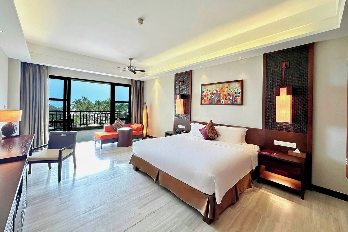 Ein modernes Hotelzimmer mit einem großen Bett, einem Schreibtisch, einem Stuhl, einem Balkon mit Blick auf Palmen und einem Deckenventilator.