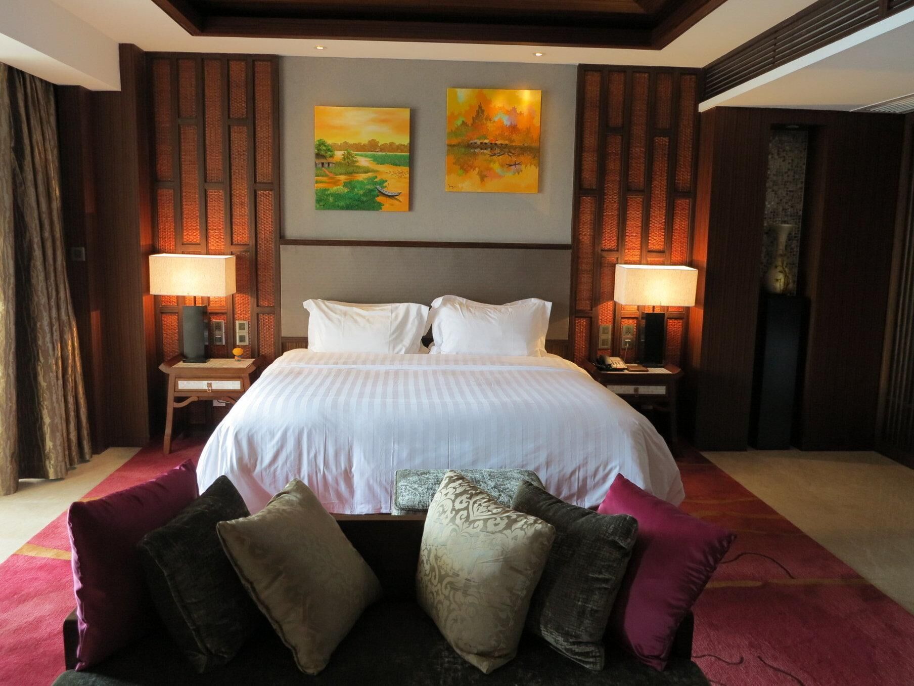 A hotel room with a large bed, two bedside tables with lamps, a red carpet, and two paintings on the wall.