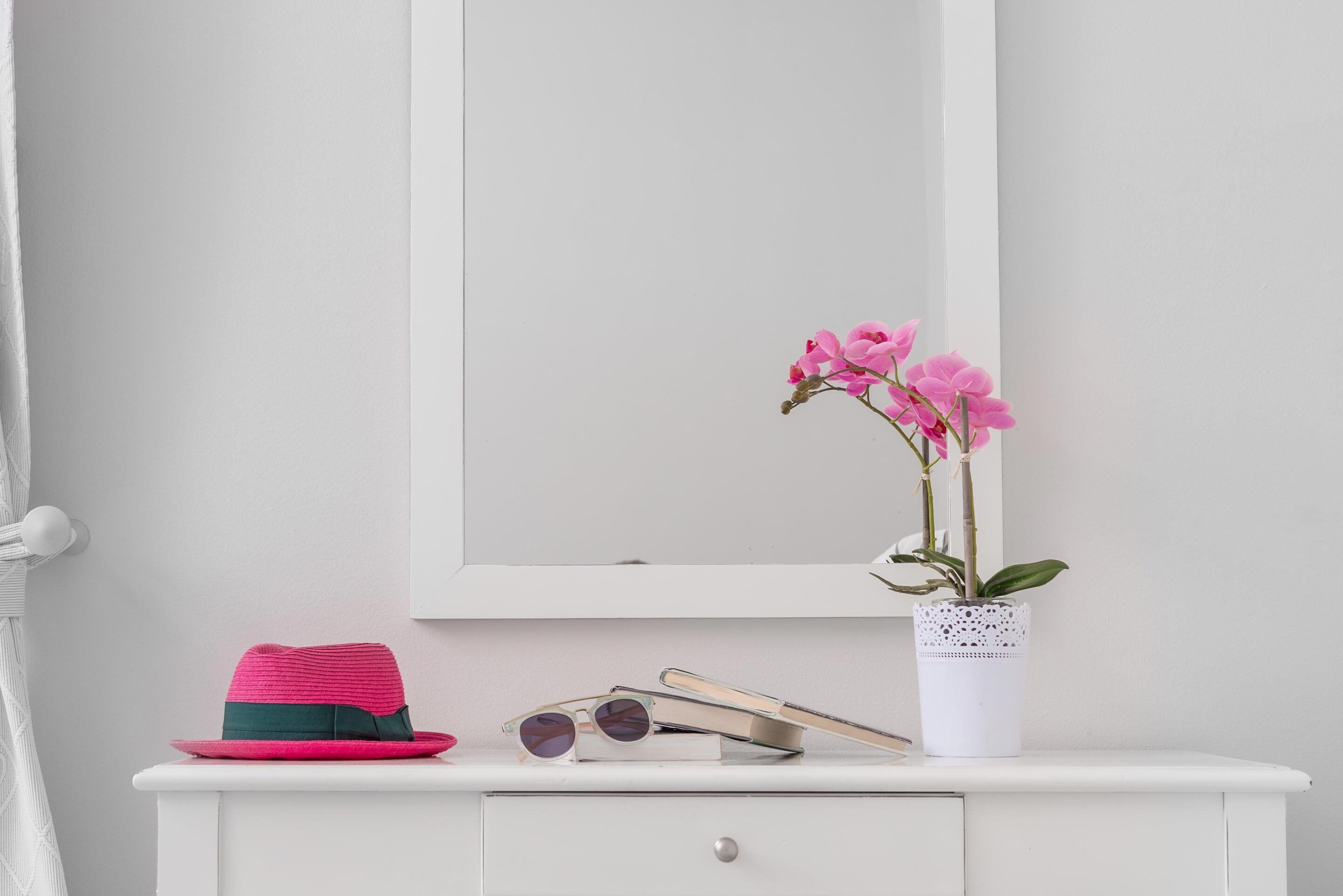 Un tocador blanco con un espejo, un sombrero rosa, gafas de sol y una orquídea en maceta.