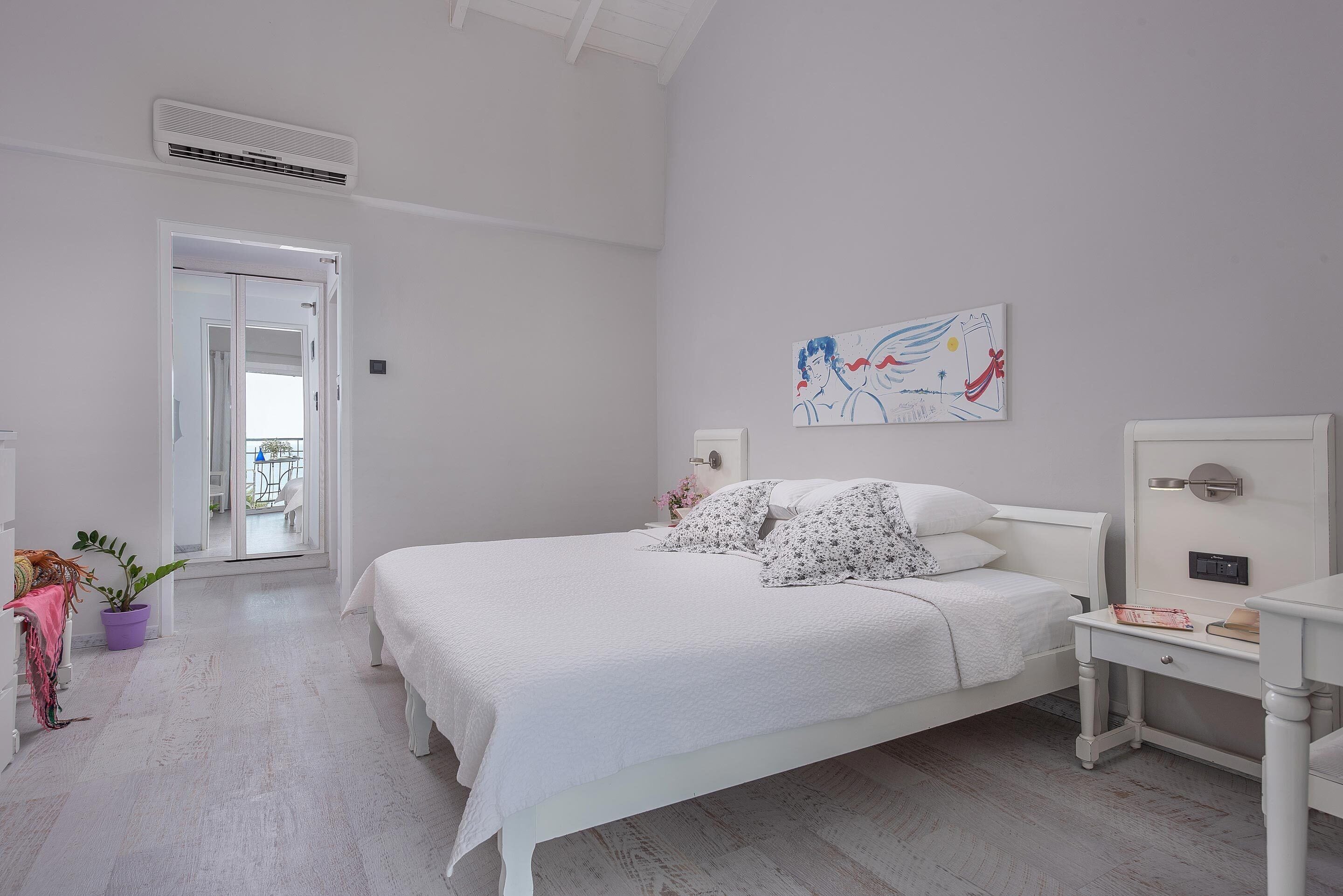 Un dormitorio con una cama blanca, una mesita de noche, un aparador, una planta en maceta y un cuadro en la pared.