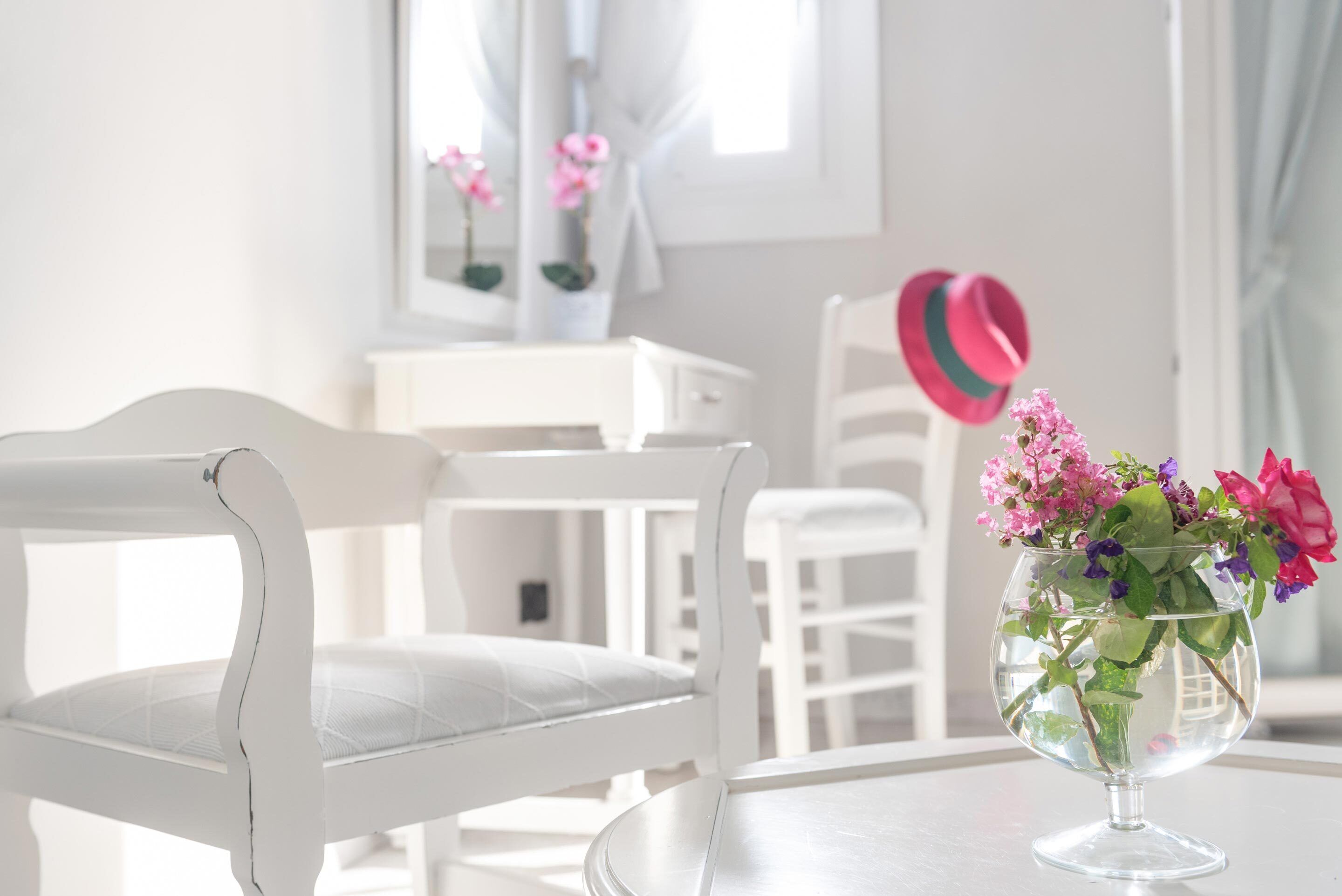 A white chair with a cushioned seat and backrest, a clear glass vase with pink and purple flowers, and a white table in a bright room with a window and curtains.