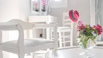 A white chair with a cushioned seat and backrest, a clear glass vase with pink and purple flowers, and a white table in a bright room with a window and curtains.
