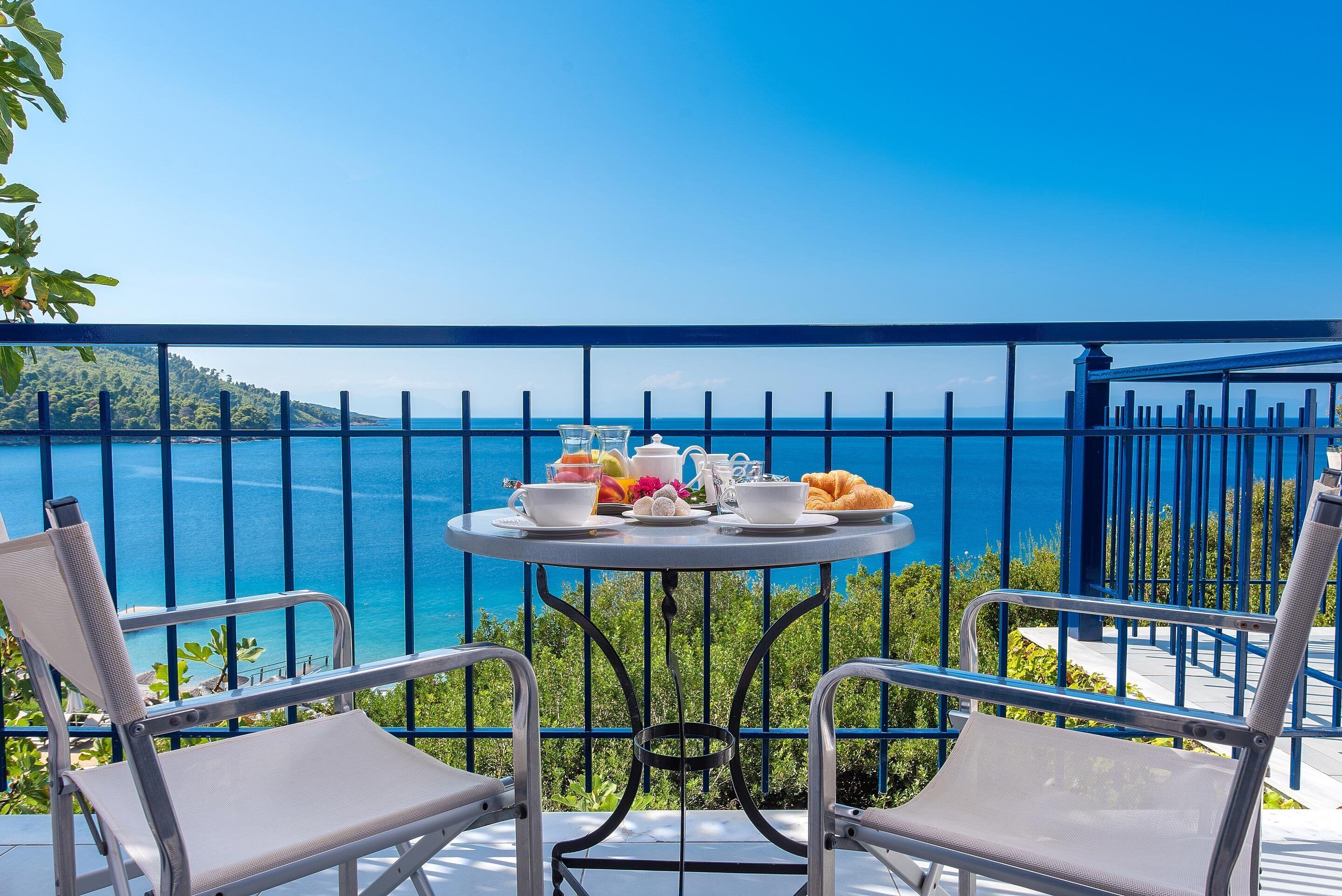 Un balcón con mesa dispuesta para el desayuno, con vista a un paisaje que incluye el mar y colinas.