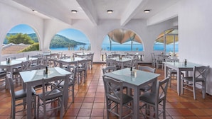 Un restaurant doté d’une terrasse, de tables et de chaises blanches, et de grandes fenêtres offrant une vue panoramique sur la mer et les collines.