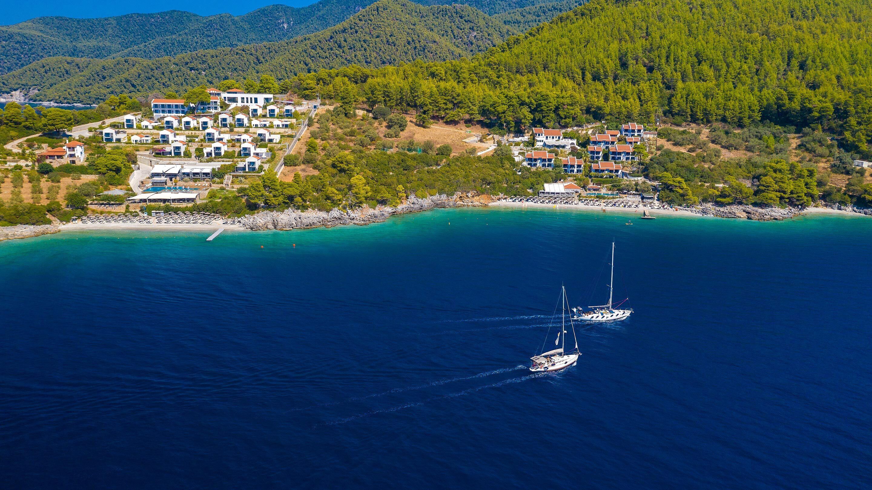 An aerial view of a coastal resort with multiple white buildings, a sandy beach, and two sailboats in the water.