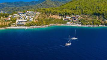 An aerial view of a coastal resort with multiple white buildings, a sandy beach, and two sailboats in the water.