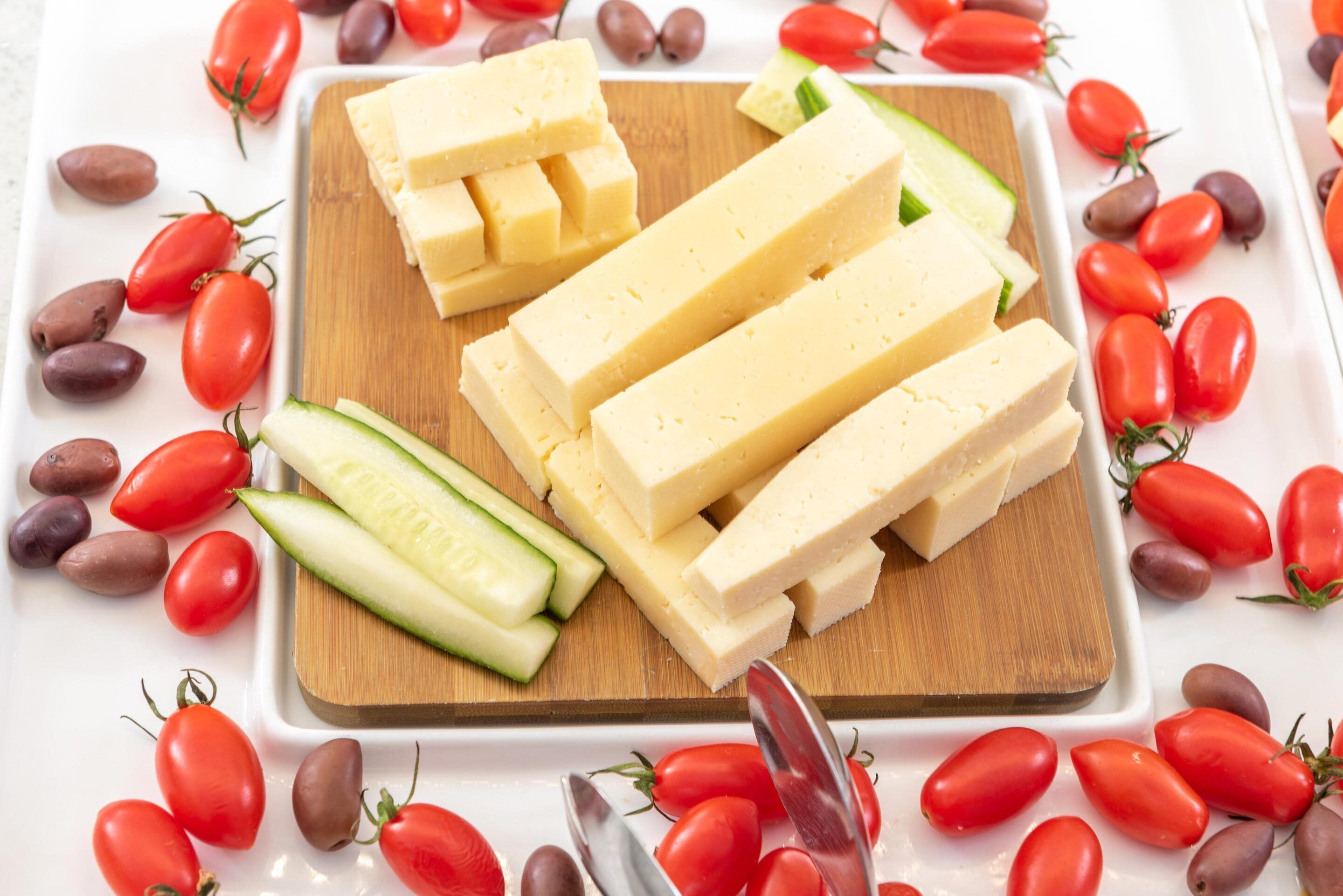 Una tabla de madera con bloques de queso y rodajas de pepino, rodeada de tomates cherry rojos y aceitunas.