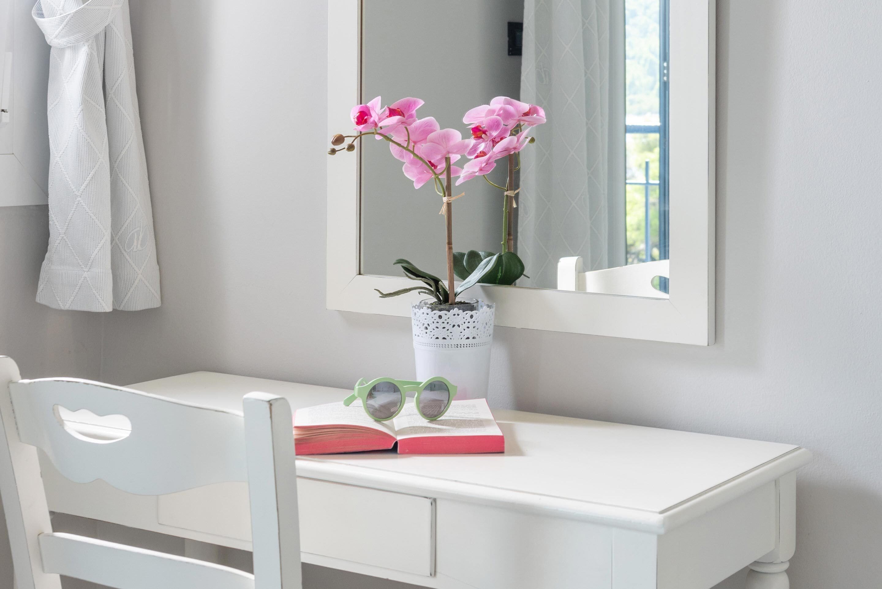 A white desk with a mirror, a vase with pink flowers, a book, and a pair of glasses.