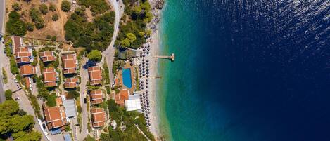 Vista aérea de un resort costero con varias cabañas, una piscina y un muelle que se adentra en el mar.