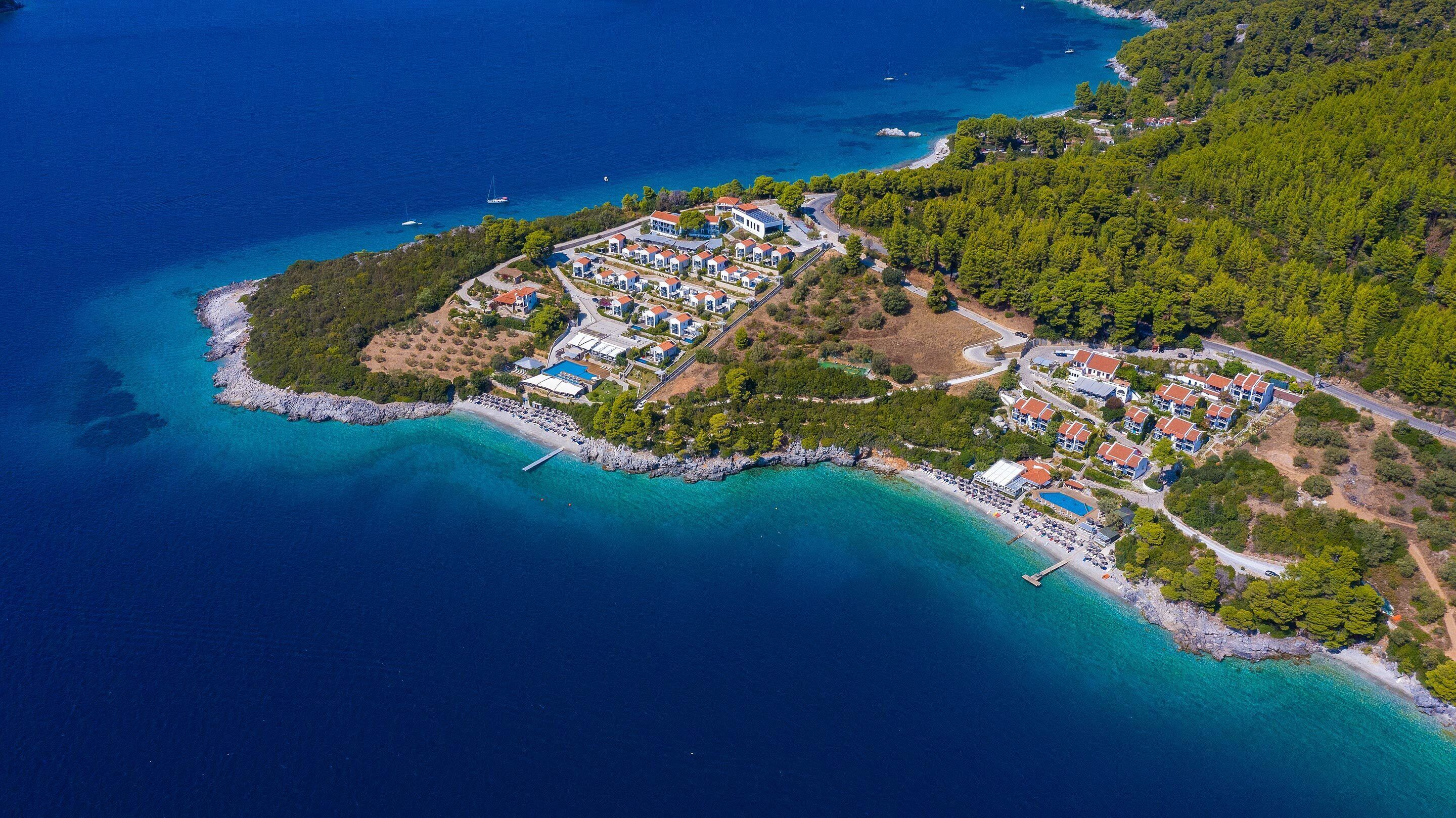 Vista aérea de un complejo turístico en la costa, con varios edificios, una playa y aguas azules cristalinas.