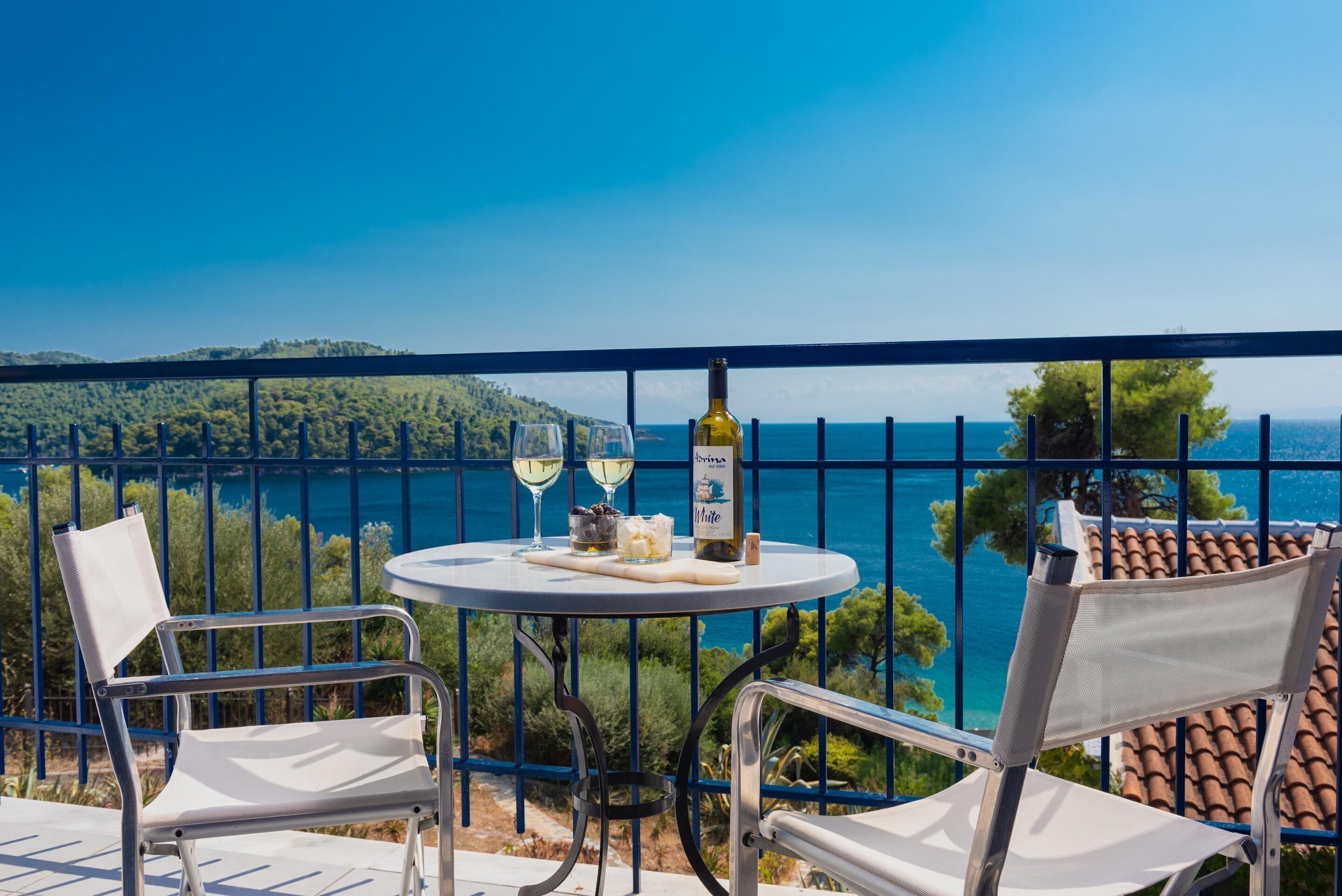 A balcony with two white chairs, a round table, a bottle of white wine, and two glasses. The view includes a coastal landscape with trees and a clear blue sky.