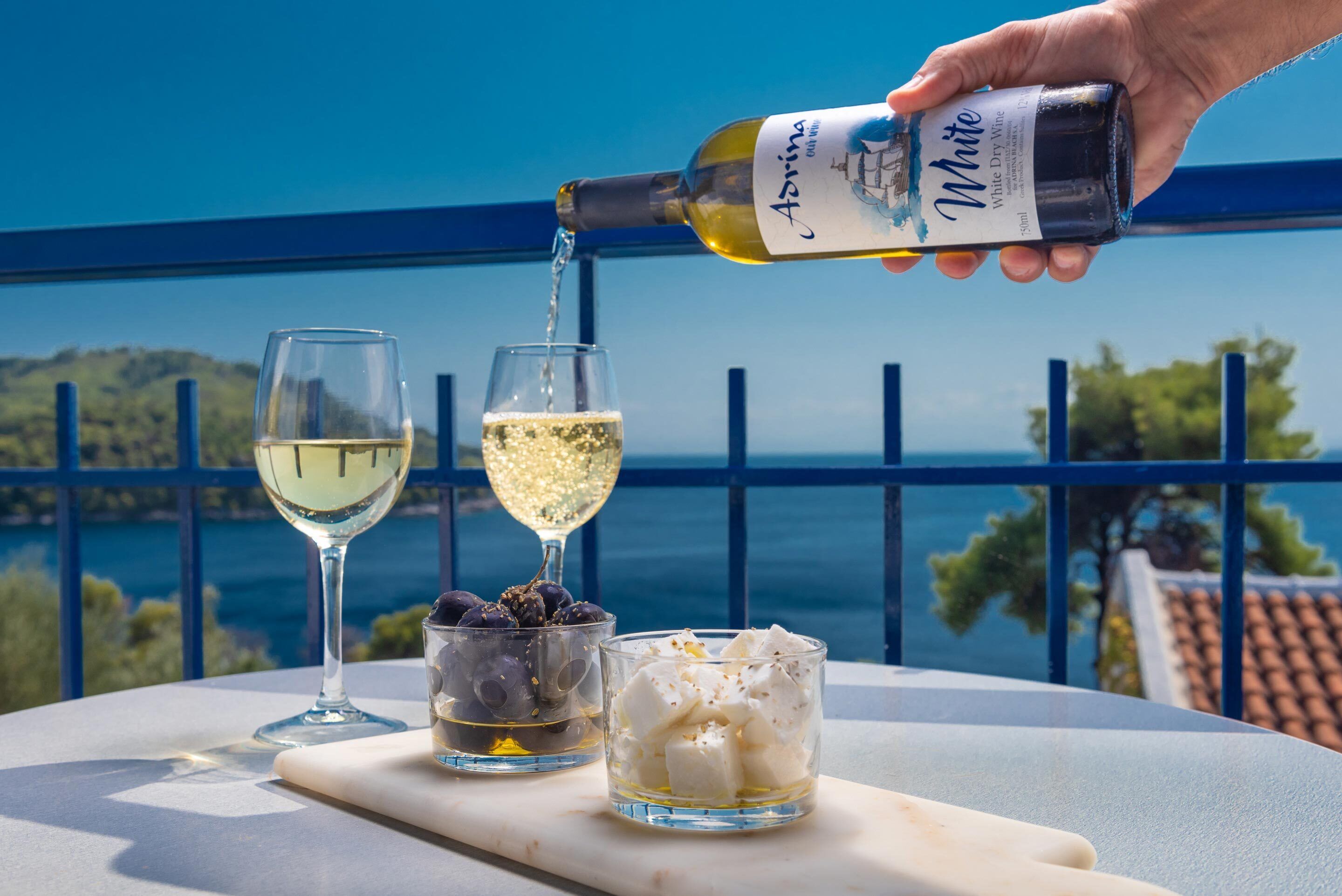Una mano sirve vino blanco en una copa, con una vista panorámica del mar y las colinas al fondo.