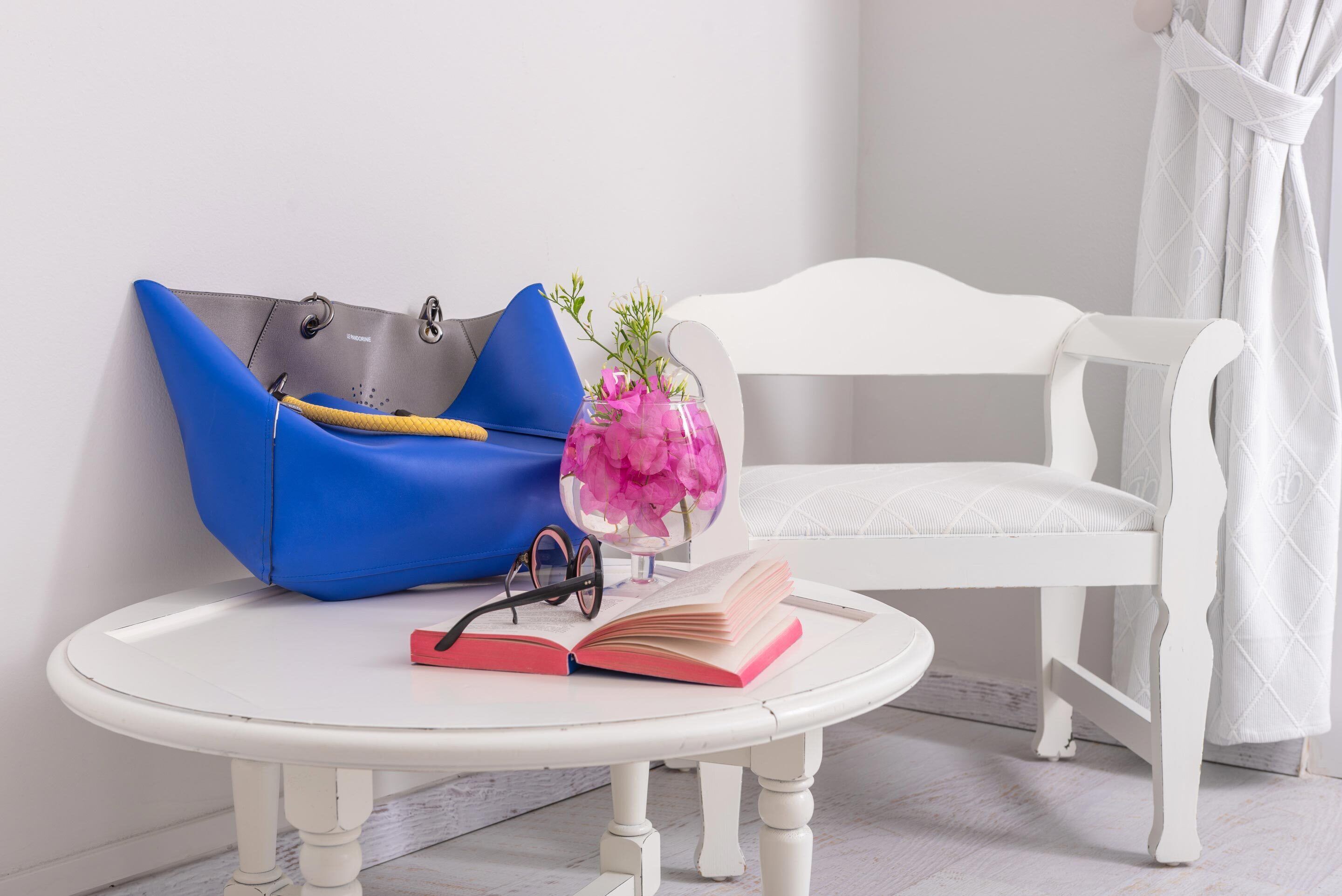 Ein weißer runder Tisch mit einer blauen Tasche, einer Brille und einer rosafarbenen Blumenvase.