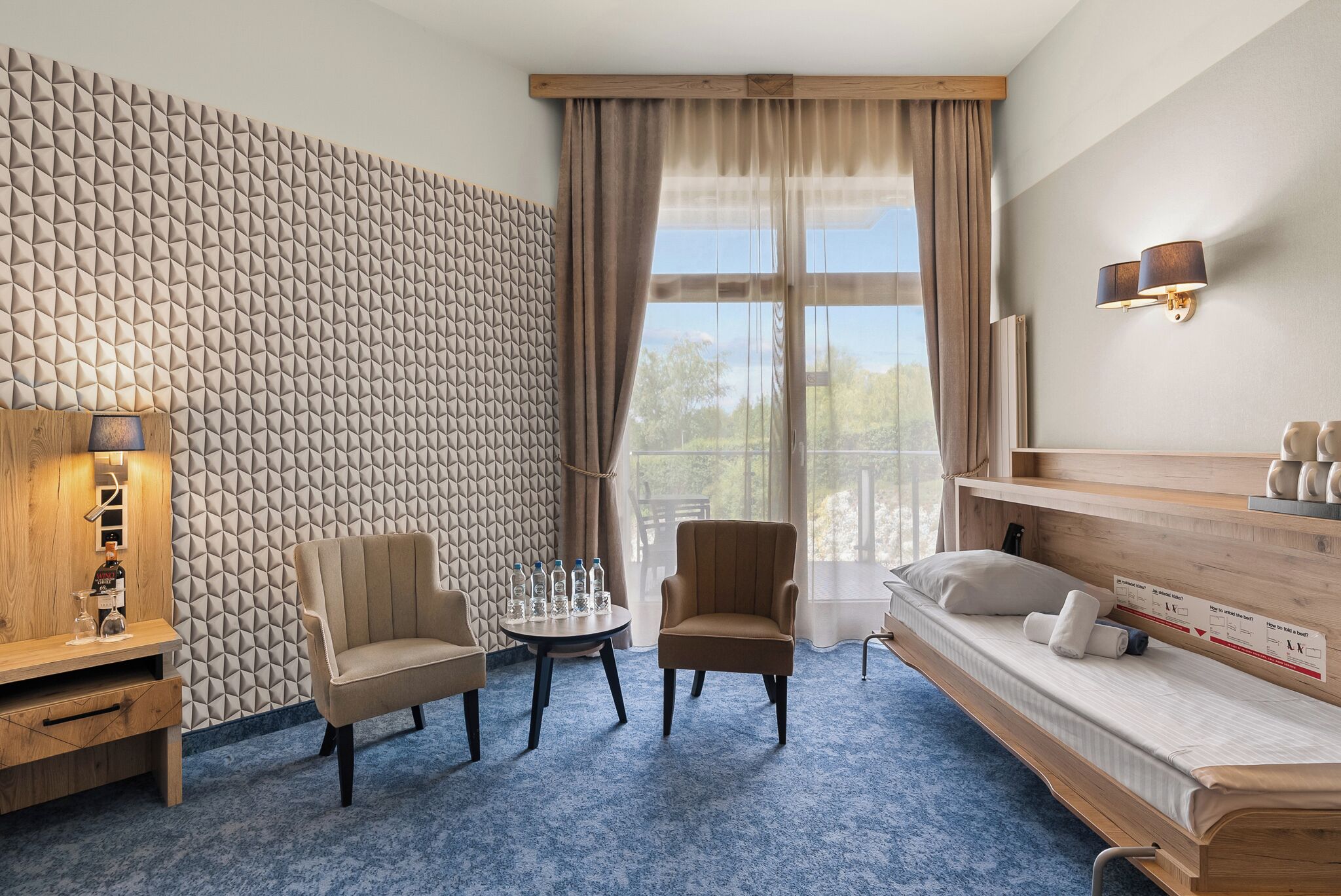 Habitación de hotel con una pared de estampado geométrico, una cama sofá, una mesita redonda con botellas y una lámpara.