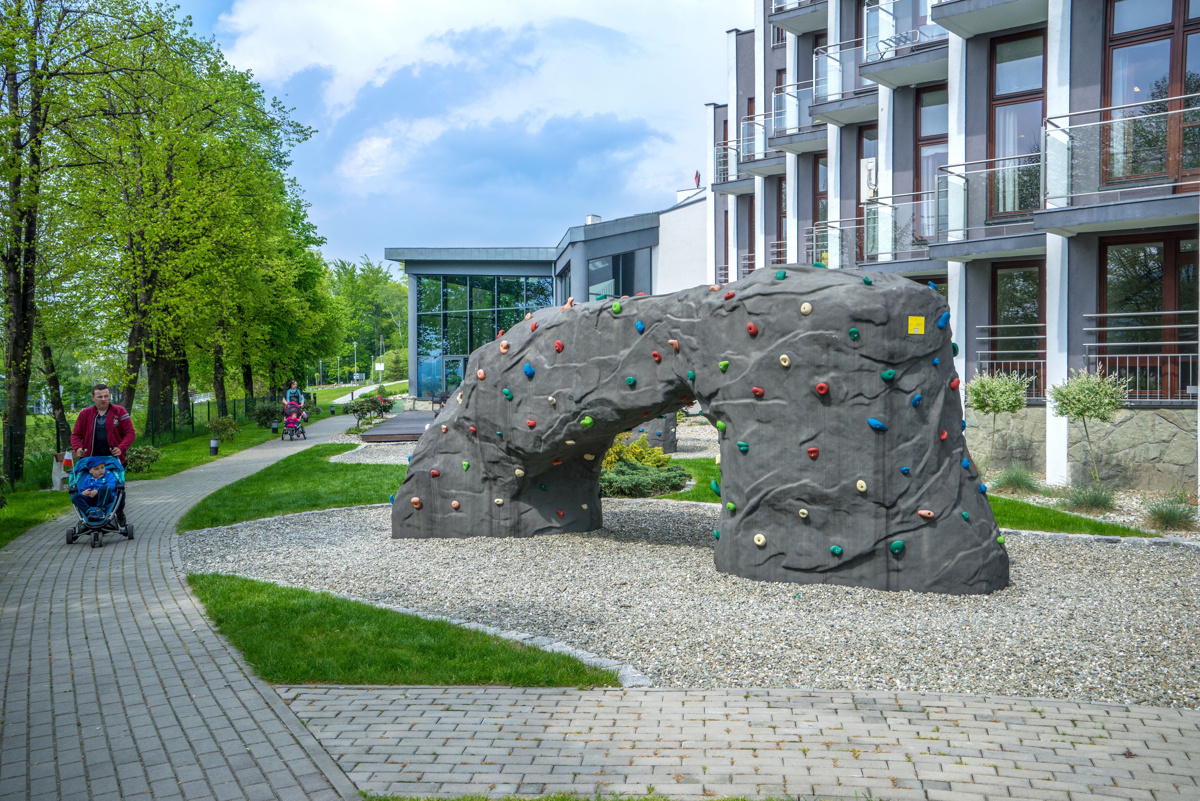 A rock climbing wall with colorful holds, a paved walkway, and a building with balconies in the background.
