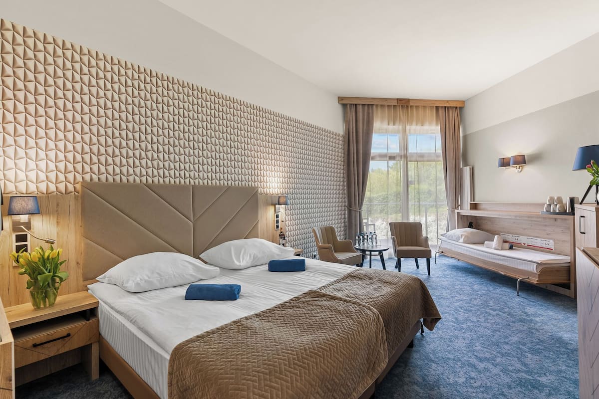 Ein Hotelzimmer mit einem großen Bett, einem Schreibtisch, einem Stuhl, einem kleinen Tisch, einem Schminktisch mit Spiegel und Blick ins Freie.