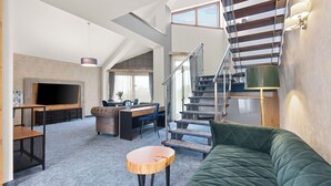 Un amplio salón con un sof á verde, una mesa de centro de madera, un televisor y una escalera con barandilla.