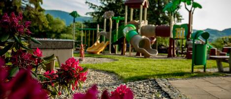A playground with slides and climbing structures, surrounded by a garden with pink flowers.