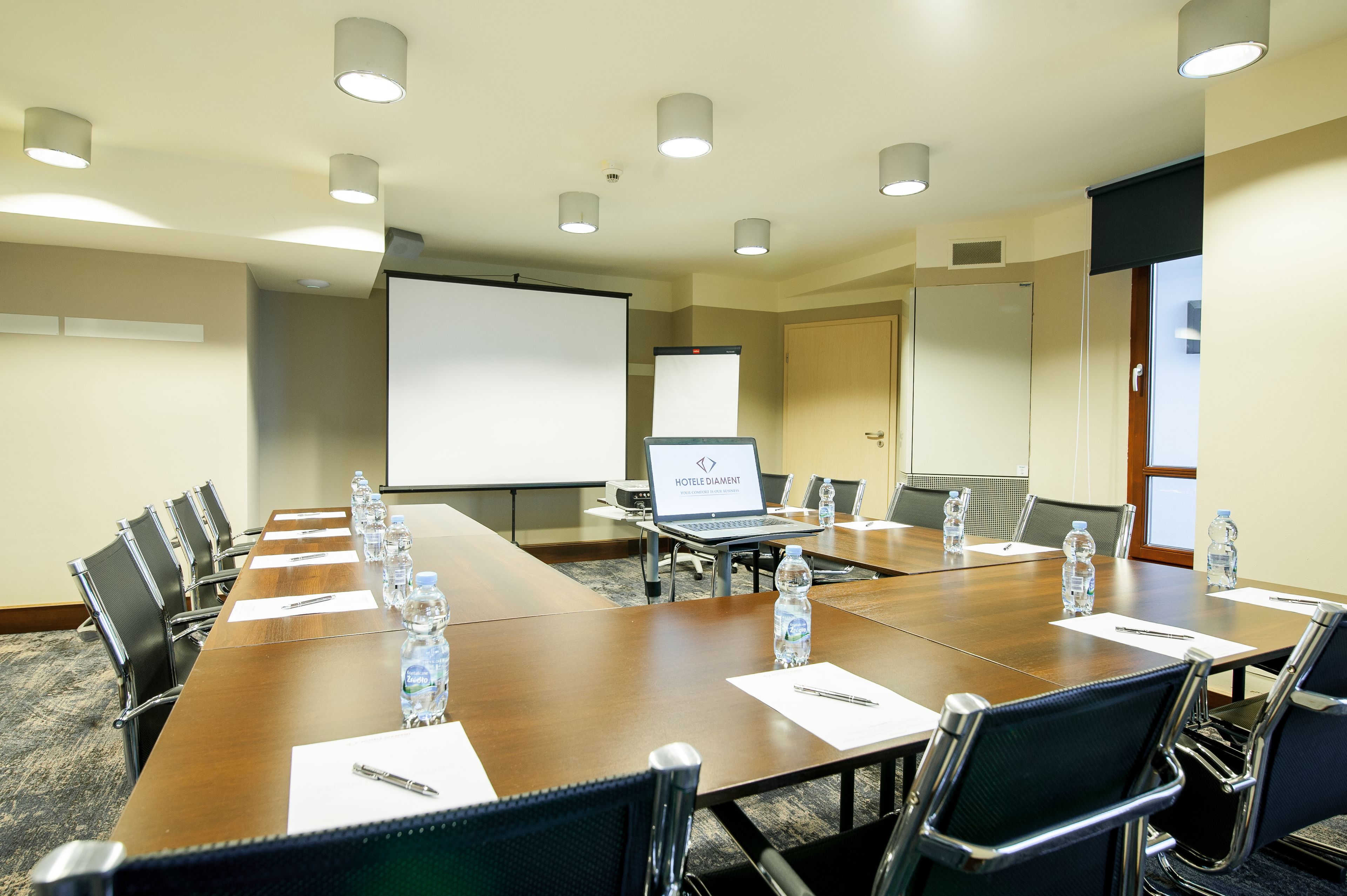 A conference room with a long table, chairs, a laptop, a projector screen, and bottled water.