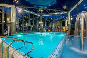 Une piscine intérieure dotée d’un toit en verre, de chaises transparentes placées au bord du bassin et d’un élément décoratif aquatique.