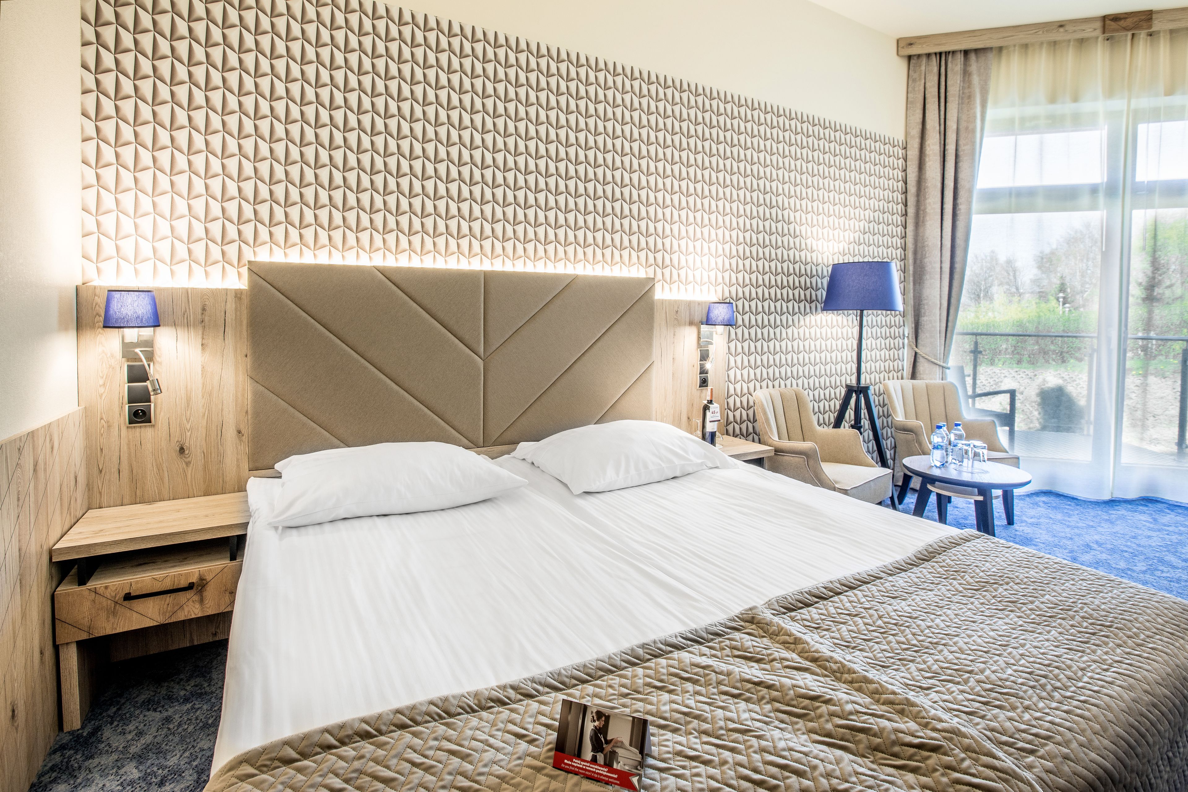 Ein Hotelzimmer mit einem großen Bett, einem Holz-Kopfteil, einer Wand mit geometrischem Muster, einem kleinen Tisch mit Stühlen und einem Fenster mit Blick.