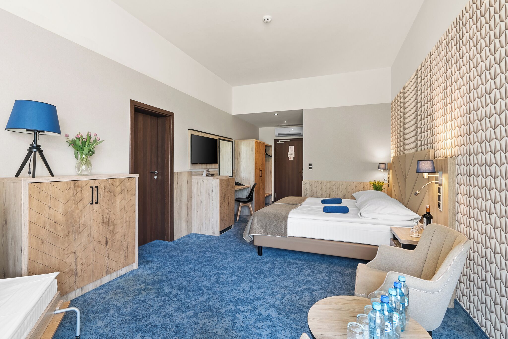 Habitación de hotel con cama, escritorio, silla, lámpara y pared con diseño geométrico.