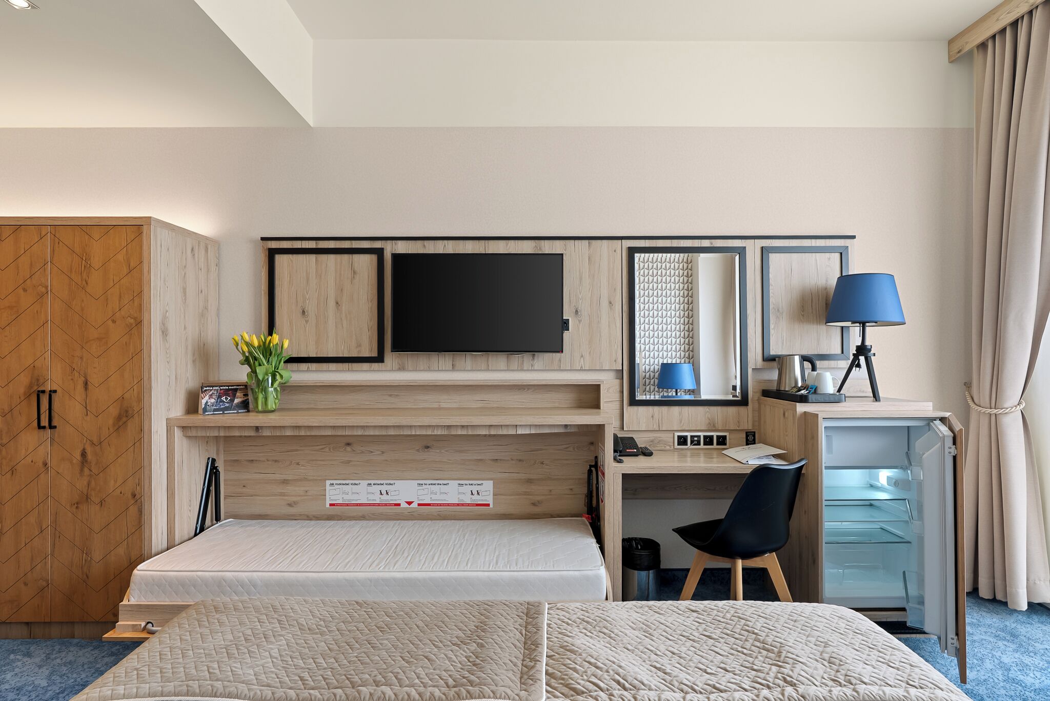 A hotel room with a bed, a desk with a computer, a TV, a lamp, and a wardrobe.