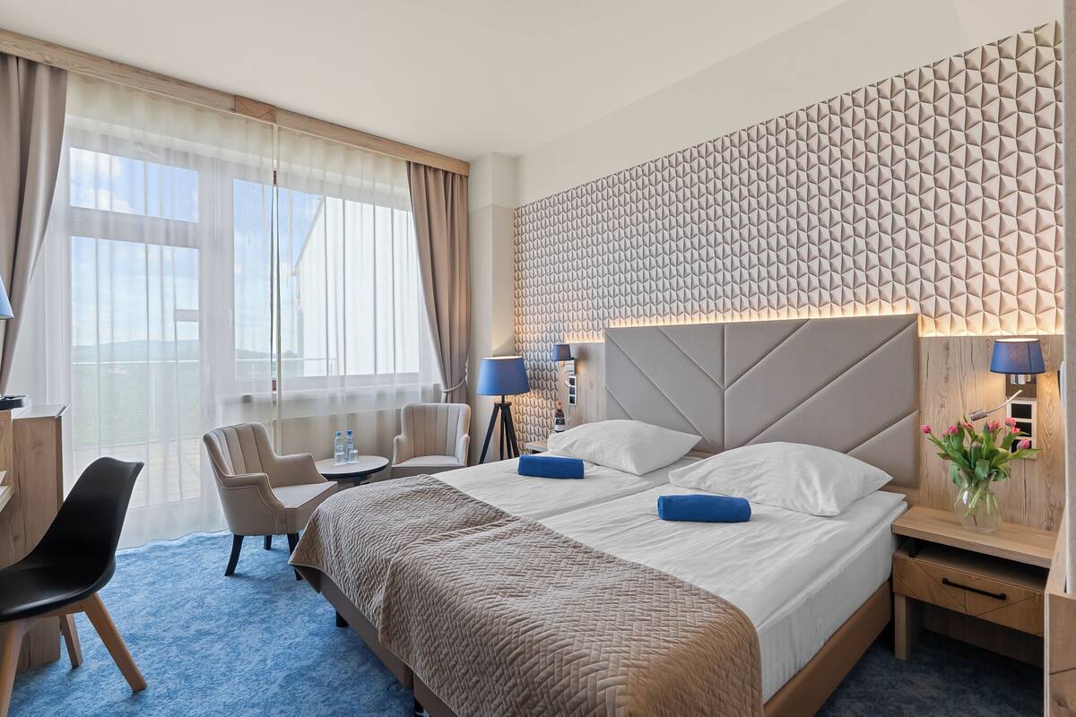 Ein Hotelzimmer mit einem großen Bett, zwei Nachttischen, einem Schreibtisch, einem Sessel und einem großen Fenster mit Vorhängen.