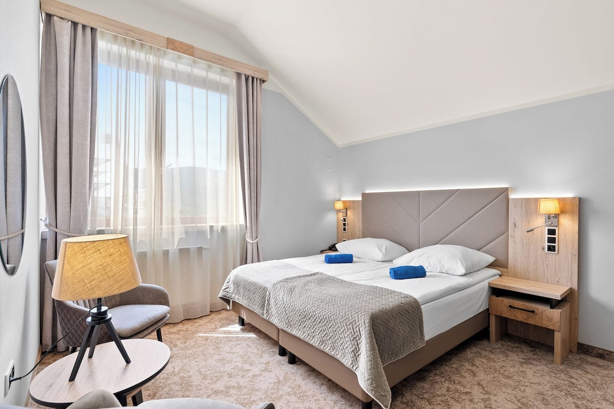 Ein Hotelzimmer mit Bett, Nachttischen, einer Lampe, einem Sessel und Blick ins Freie.