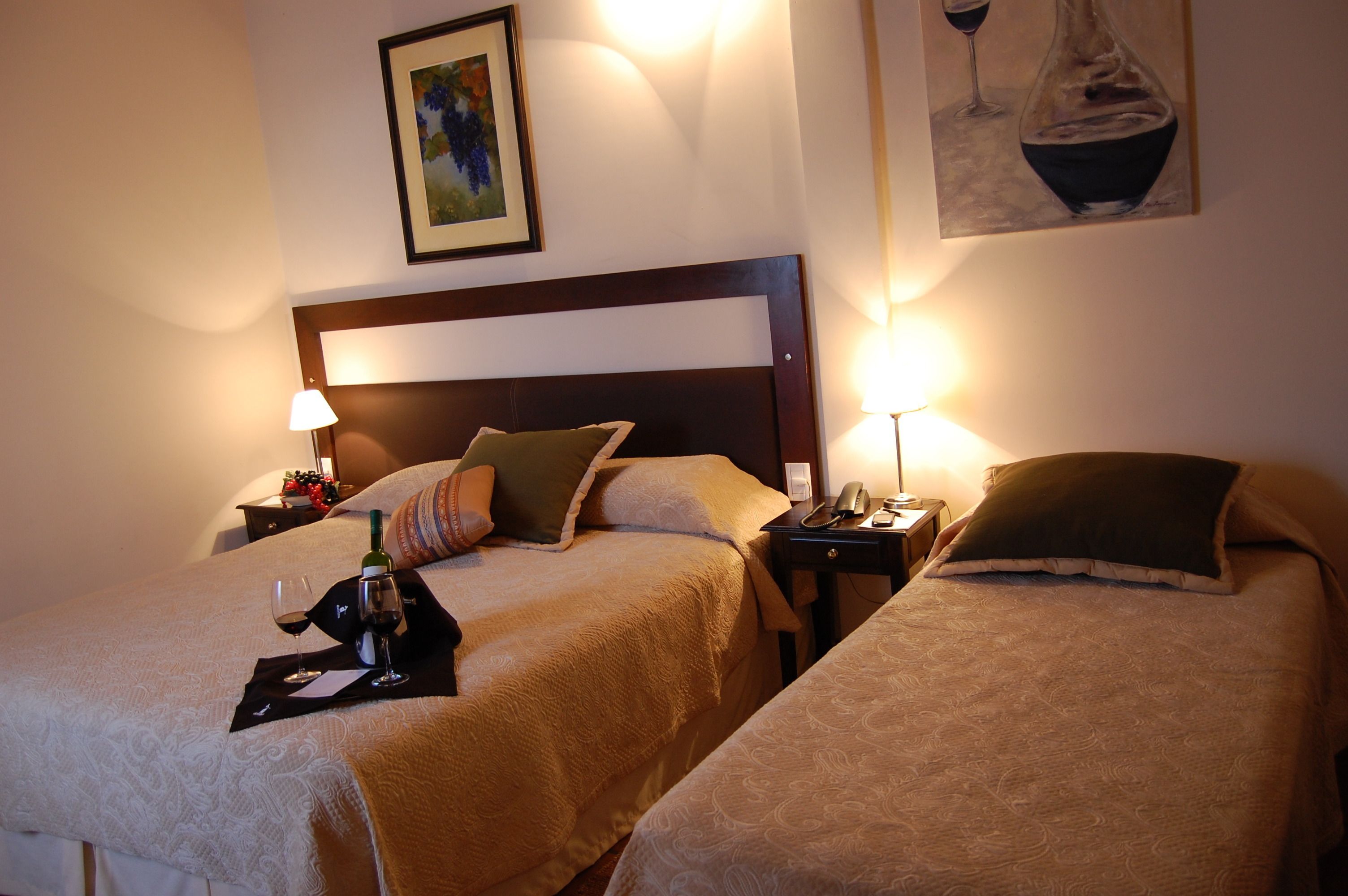 Habitación de hotel con dos camas, cada una con una almohada, una mesita de noche con una lámpara y una botella de vino sobre la cama de la izquierda.