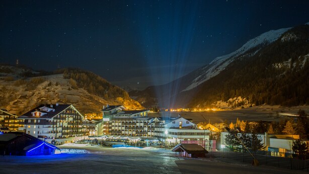 Eine verschneite Bergstadt bei Nacht mit beleuchteten Gebäuden und einer Seilbahn.